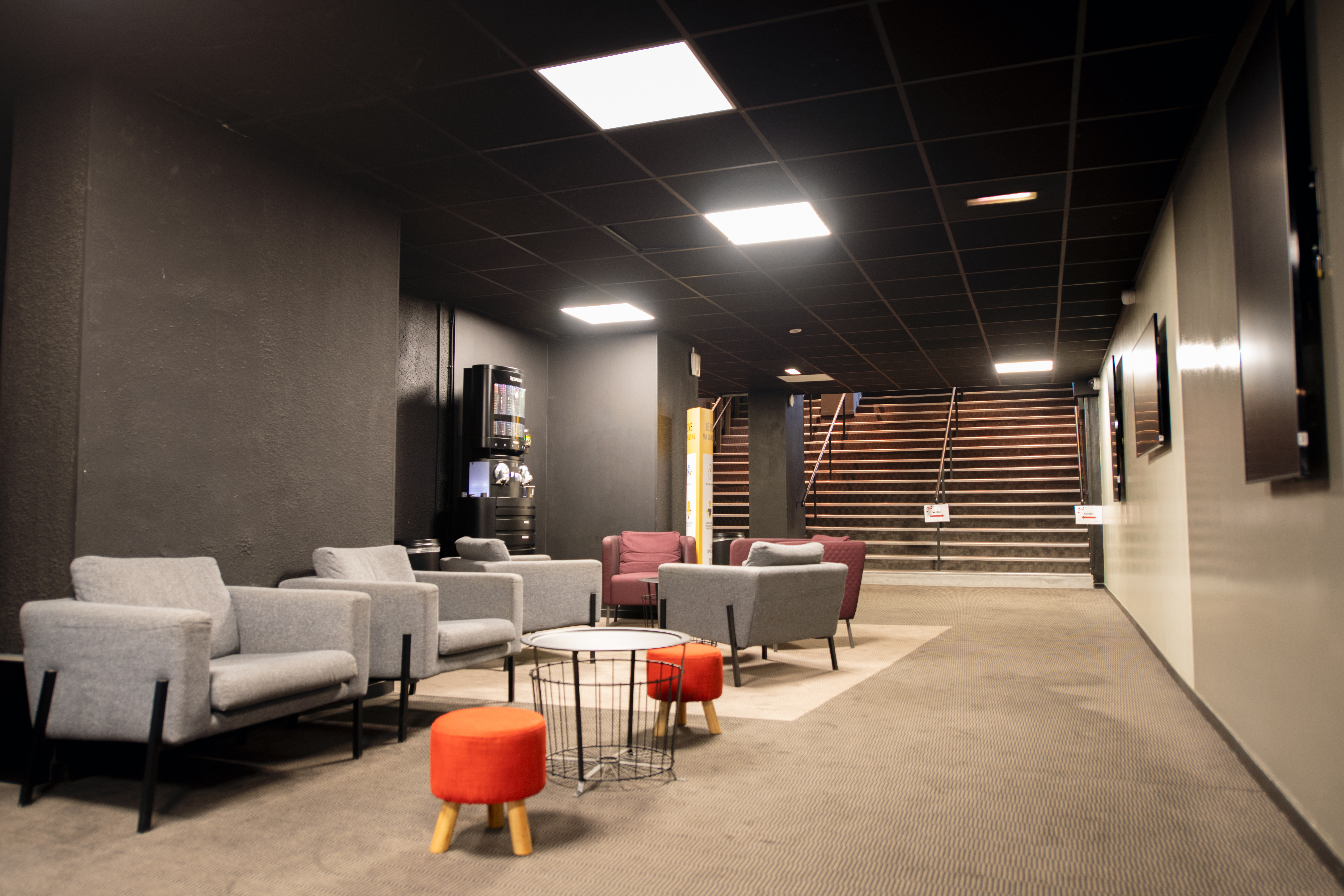 Espace de réception du cinéma avec fauteuils gris et rouges, tables basses et machine à café, près d’un escalier.