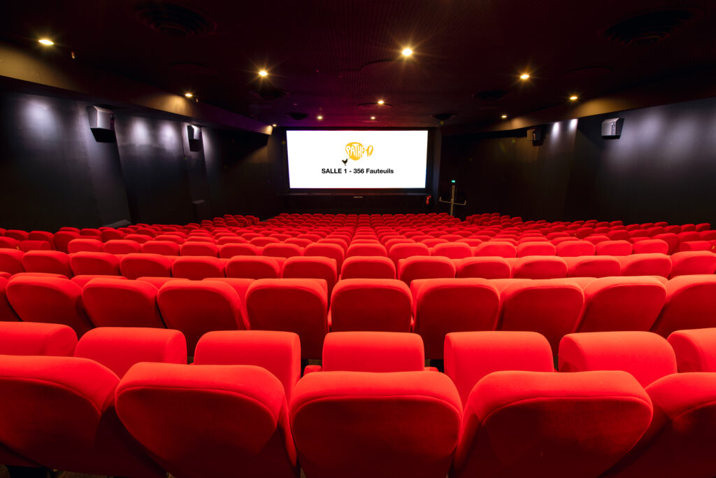 Vue depuis l’arrière de la salle 1 du cinéma Pathé Comédie avec rangées de fauteuils en velours rouges orientés vers l’écran.