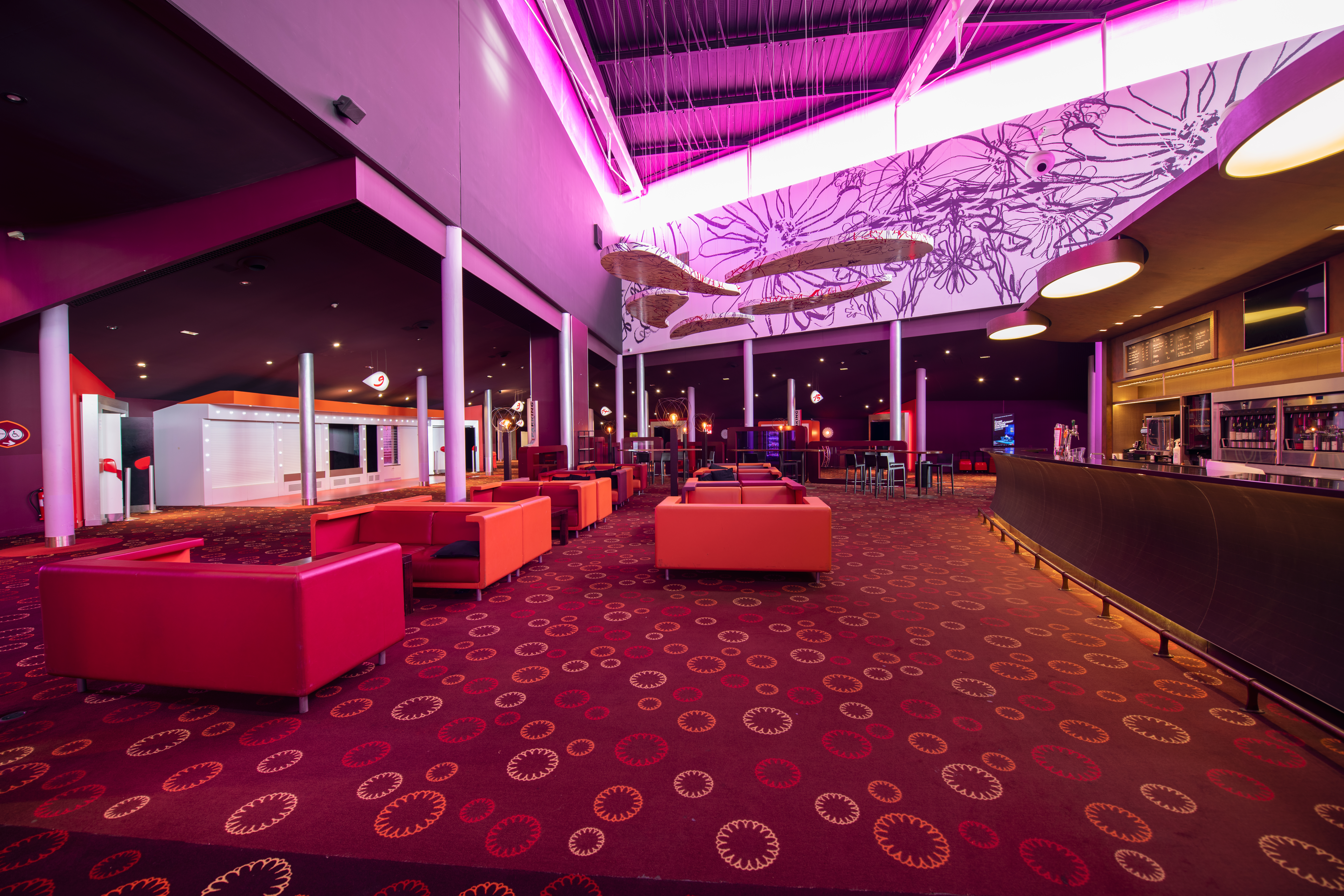 Espace lounge du cinéma avec mobilier coloré, décorations suspendues et éclairage rose immersif.