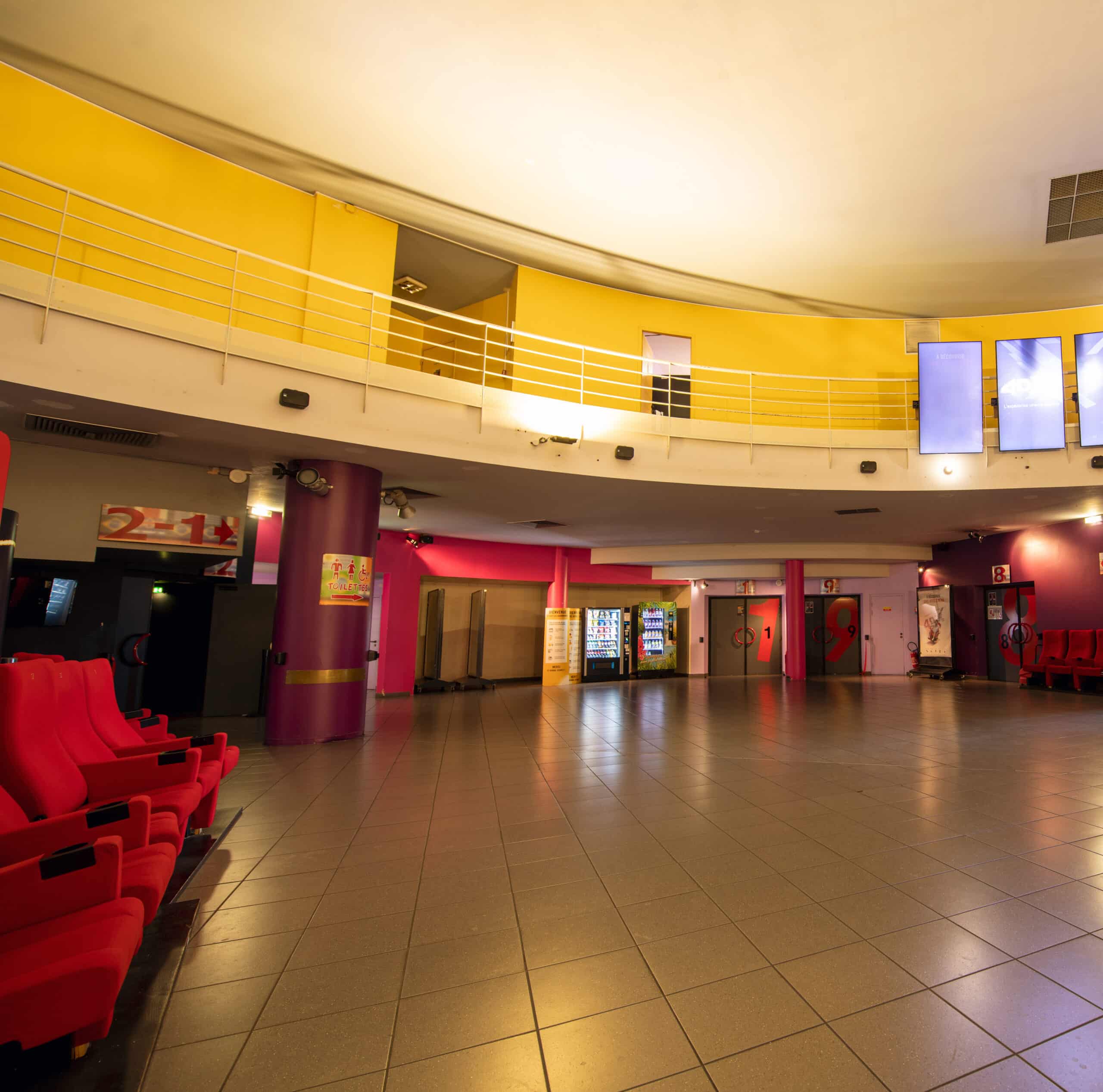 Hall spacieux aux couleurs vives avec murs jaunes et roses, sièges rouges, distributeurs automatiques et numéros indiquant les salles.