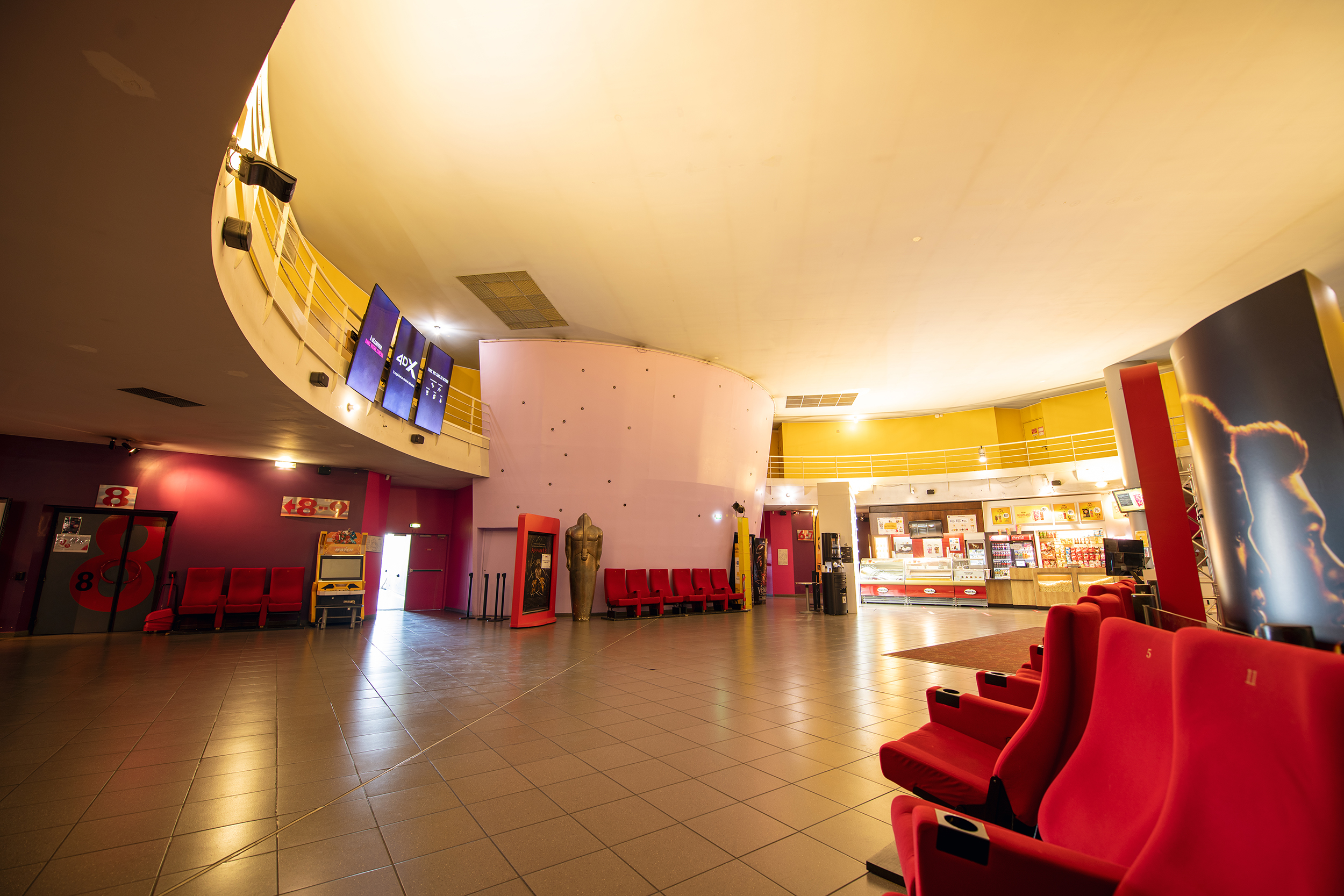 Hall spacieux aux couleurs vives avec murs jaunes et roses, sièges rouges, distributeurs automatiques et numéros indiquant les salles.