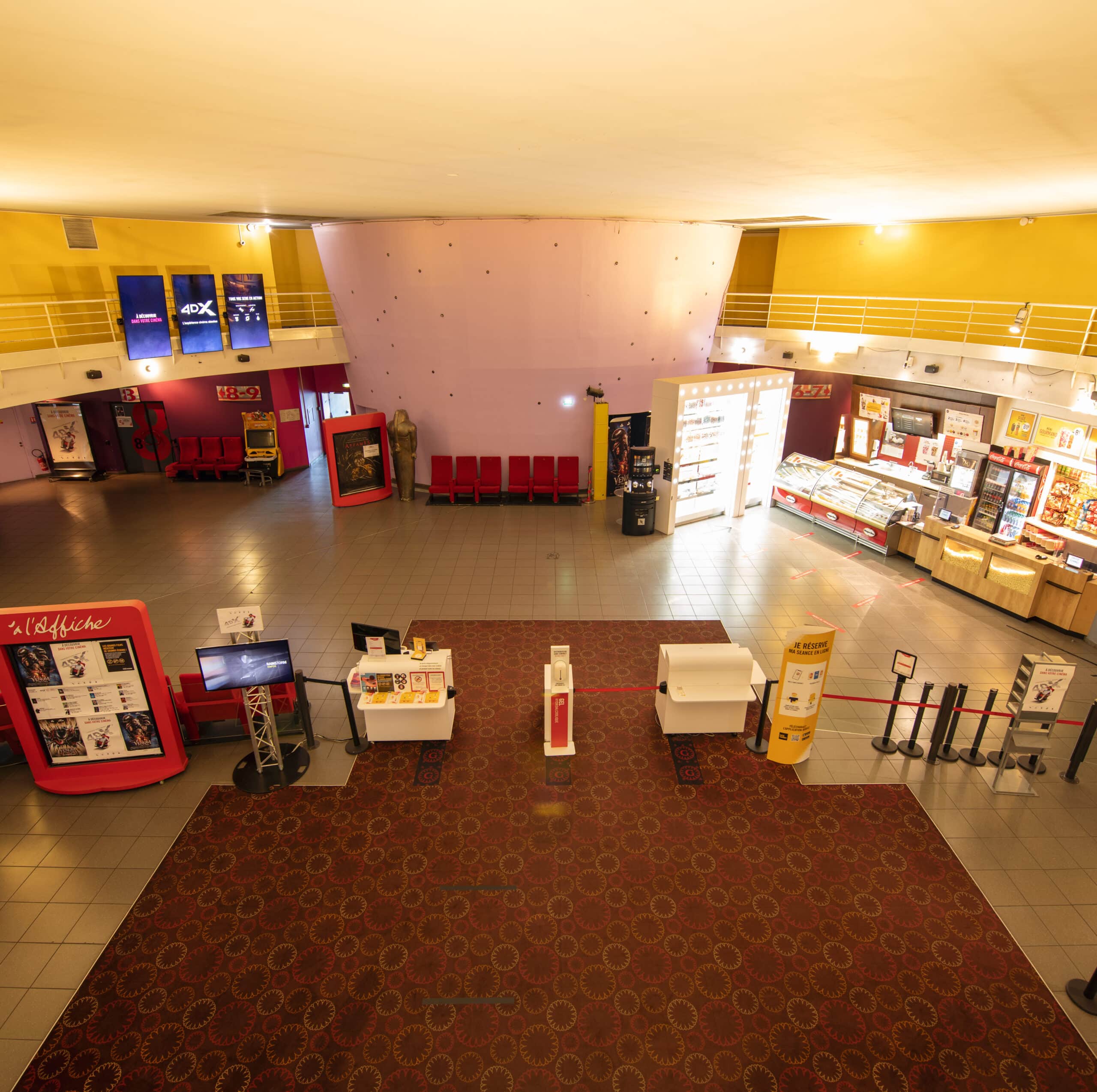 Vue d'ensemble d'un hall de cinéma avec comptoirs de vente, distributeurs automatiques, affichages numériques et sièges rouges.
