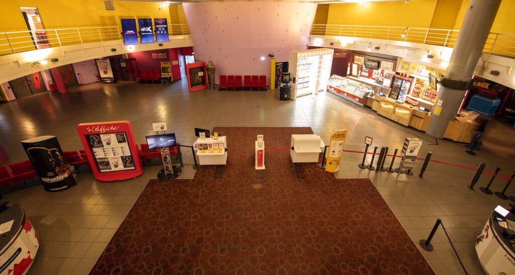 Hall spacieux aux couleurs vives avec murs jaunes et roses, sièges rouges, distributeurs automatiques et numéros indiquant les salles.