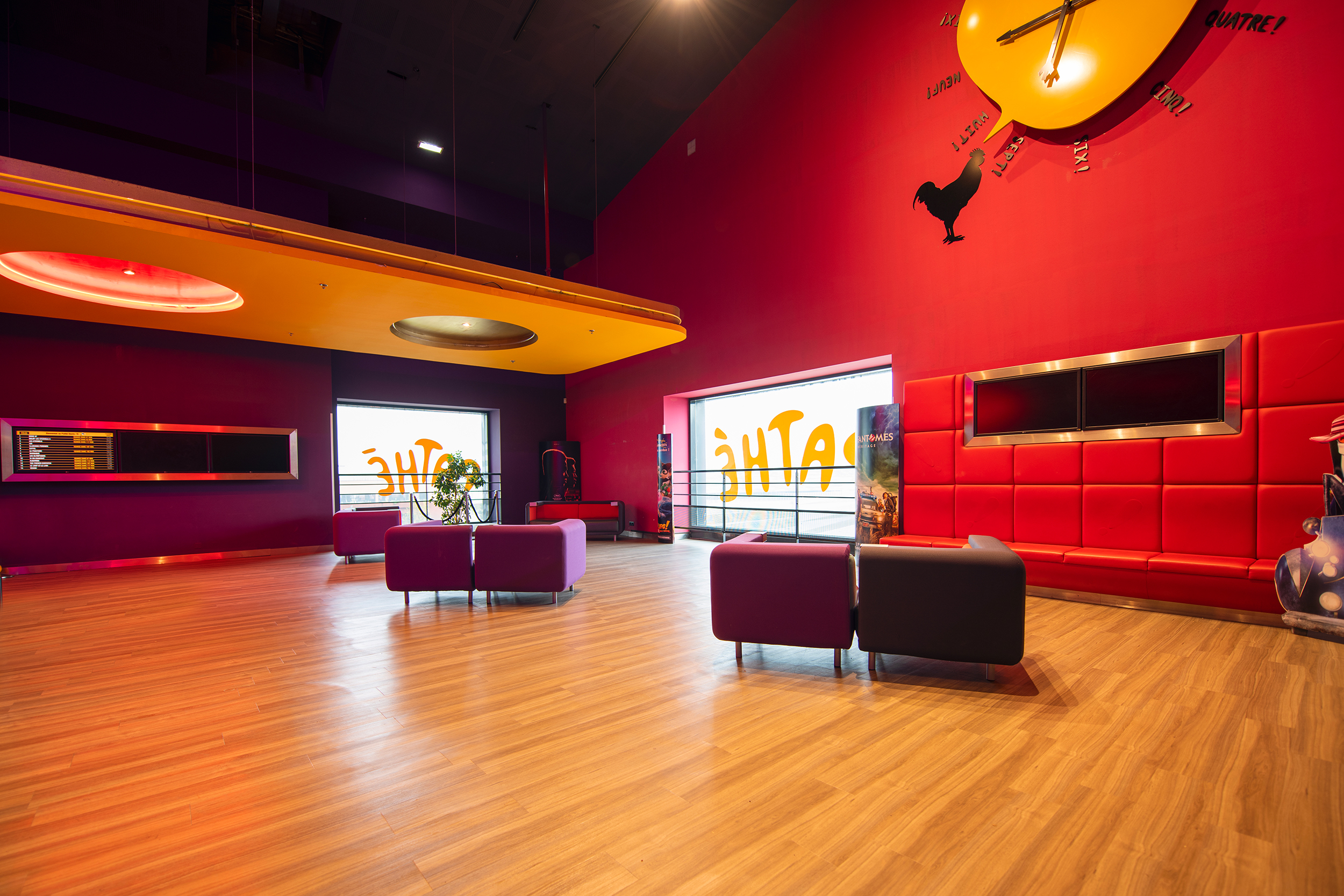Espace coloré dans un cinéma avec des murs rouges et violets, un plafond jaune, des fauteuils modernes, et un sol en bois clair.