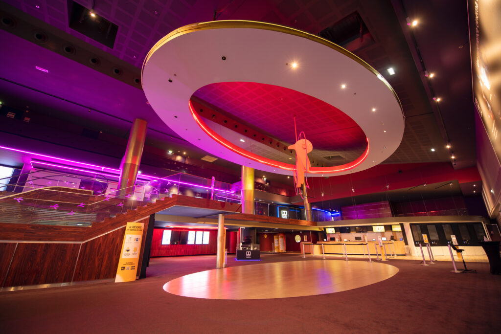 Grand hall intérieur coloré avec un plafond orné d’un cercle lumineux rouge et doré suspendu. En dessous, un escalier en bois mène à une mezzanine avec des rampes en verre et des lumières violettes. Le sol en moquette brune entoure une zone circulaire claire. À droite, un comptoir de billetterie est visible.