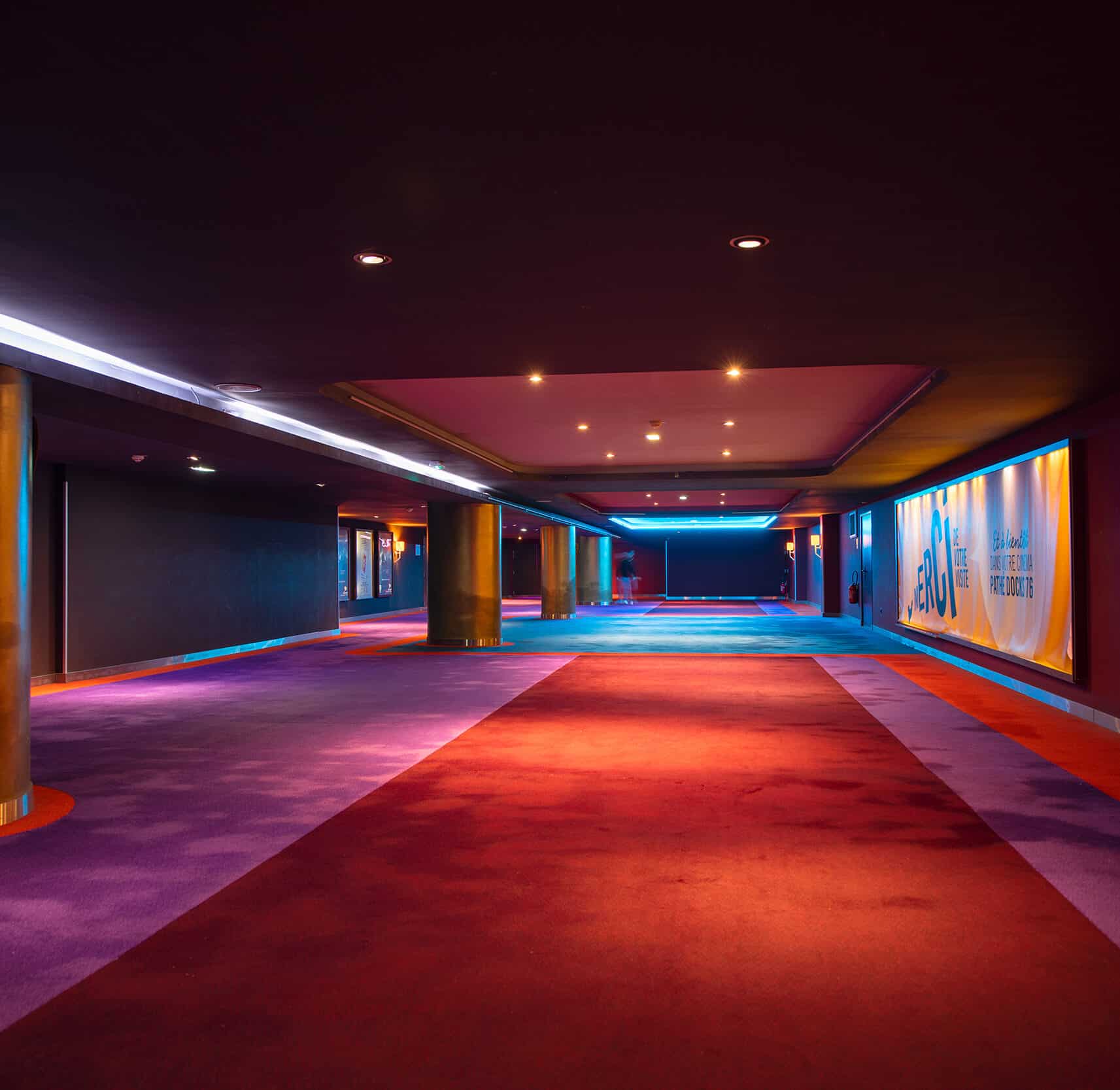 Couloir spacieux et moderne dans un cinéma avec un éclairage tamisé, tapis colorés rouge et violet, et des poteaux métalliques dorés.