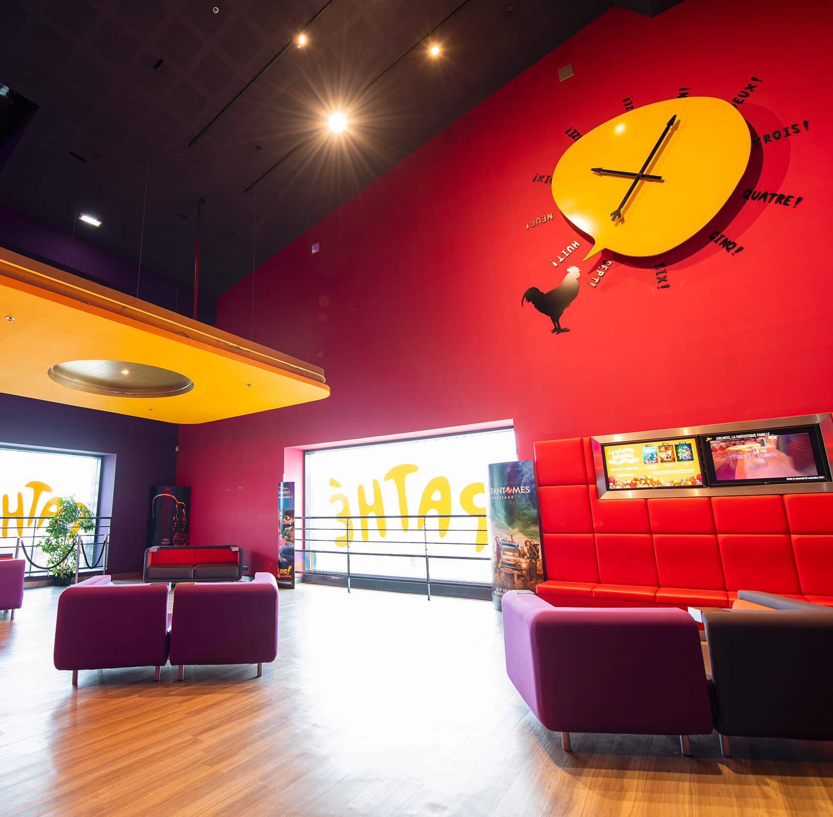 Espace coloré dans un cinéma avec des murs rouges et violets, un plafond jaune, des fauteuils modernes, et un sol en bois clair.