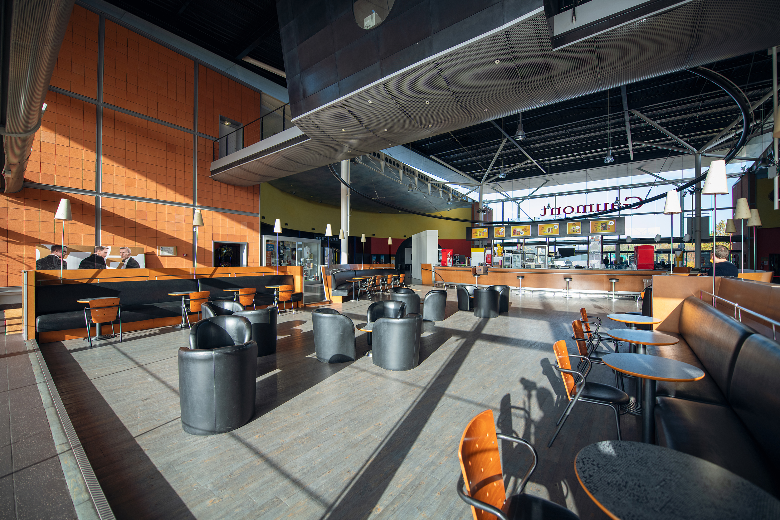 Espace café spacieux et lumineux avec des fauteuils noirs, des tables en bois, un mur orange, et une grande baie vitrée laissant entrer la lumière naturelle.