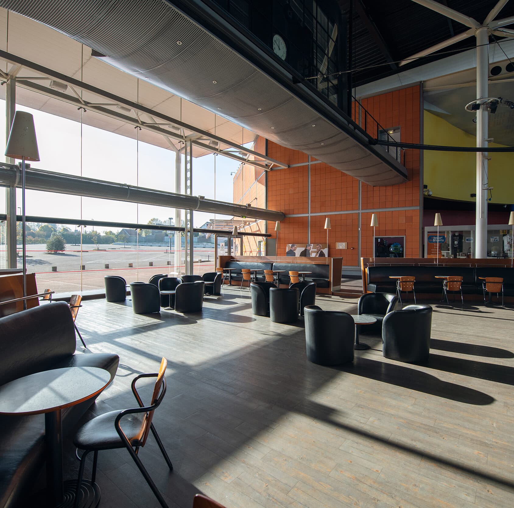 Espace café spacieux et lumineux avec des fauteuils noirs, des tables en bois, un mur orange, et une grande baie vitrée laissant entrer la lumière naturelle.