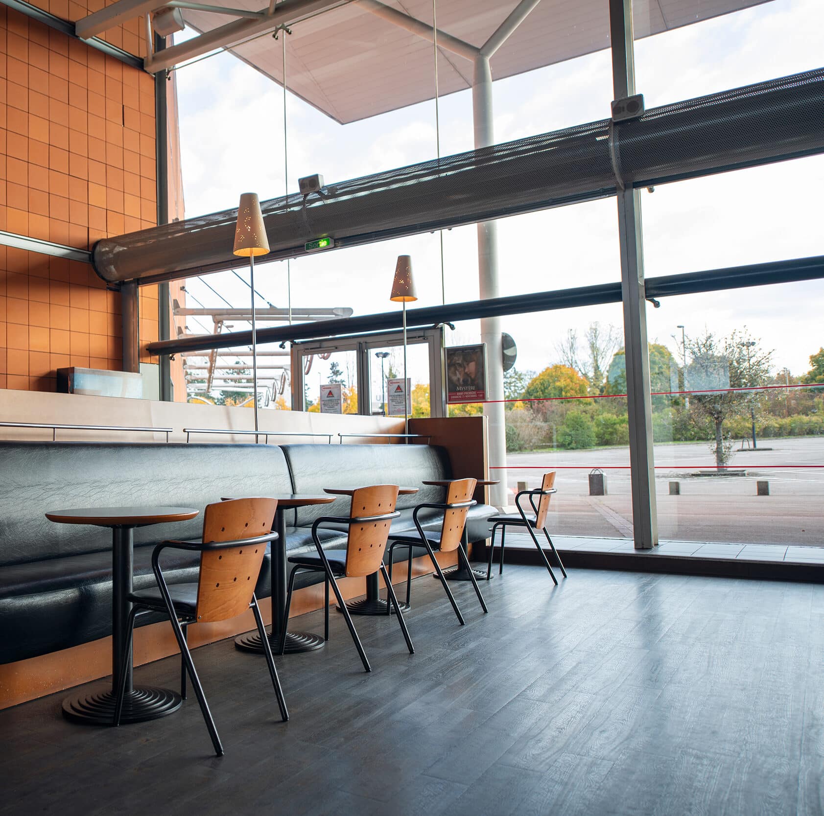 Espace café lumineux dans un cinéma, avec des chaises en bois, des tables rondes, un banc rembourré et une grande baie vitrée offrant une vue sur l'extérieur.