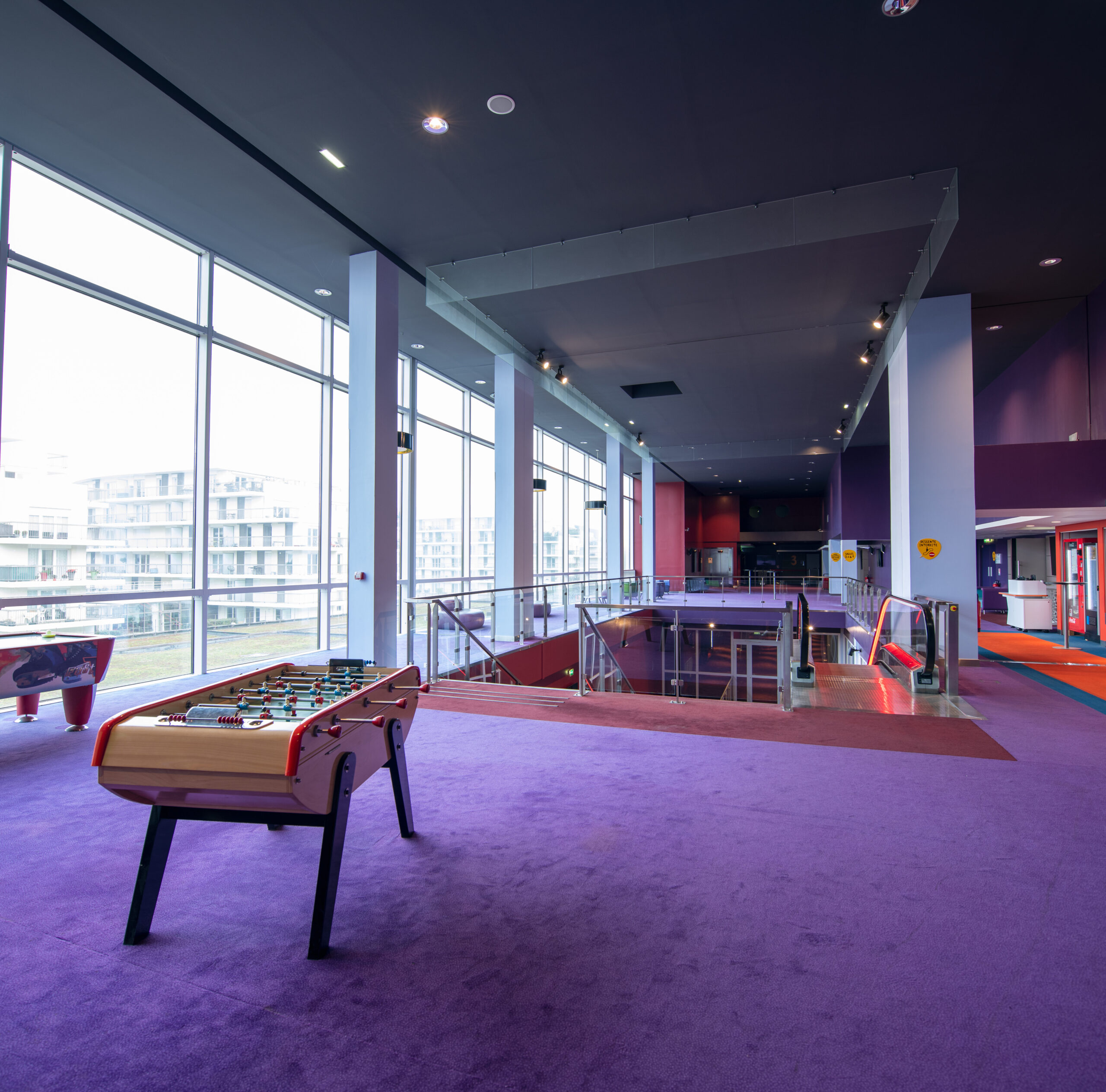 Étage lumineux dans un cinéma avec un tapis violet, une grande baie vitrée, un baby-foot et des escaliers menant aux niveaux inférieurs.