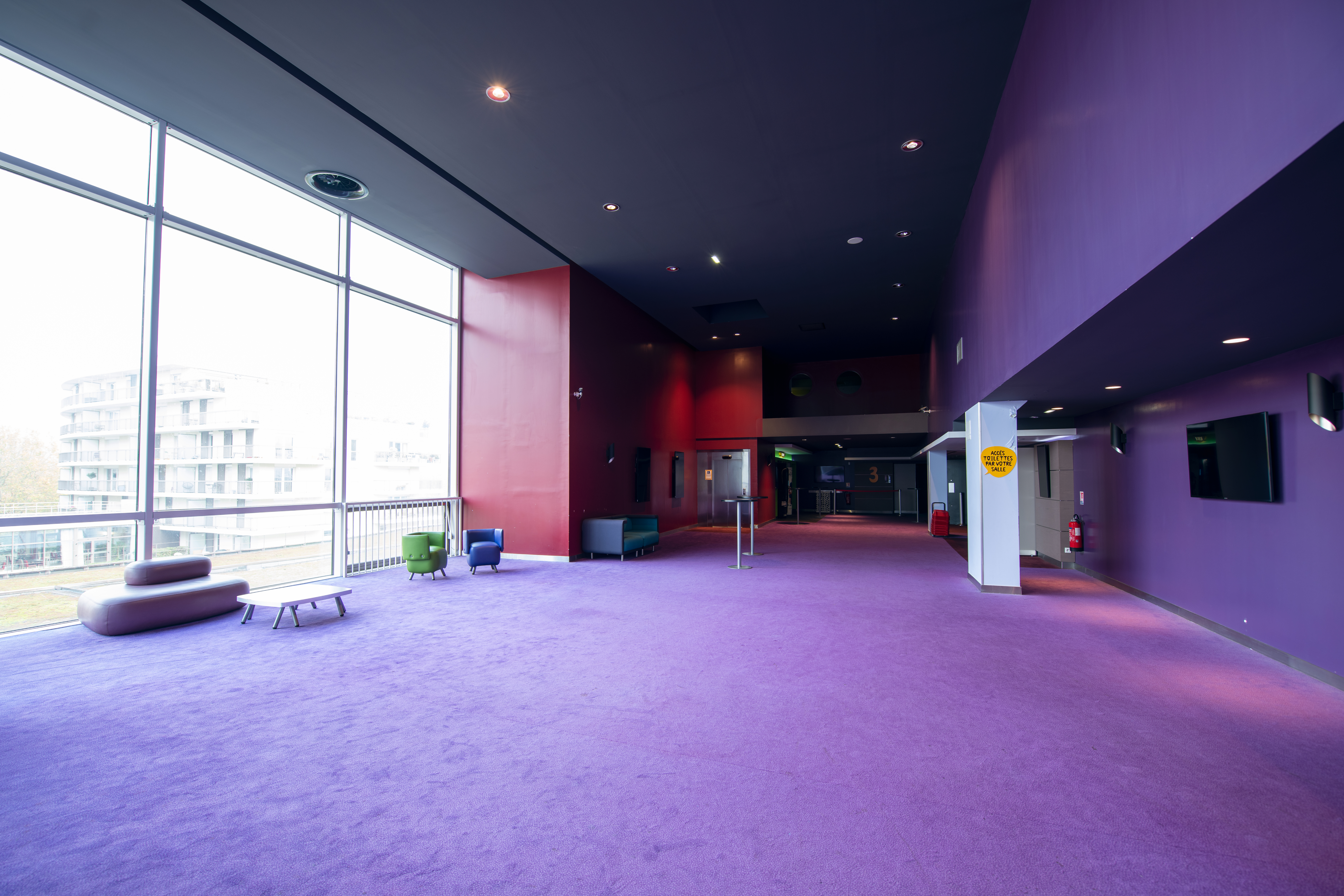 Espace lumineux avec un tapis violet, des fauteuils colorés, une grande baie vitrée donnant sur l'extérieur, et des accès aux salles de cinéma.