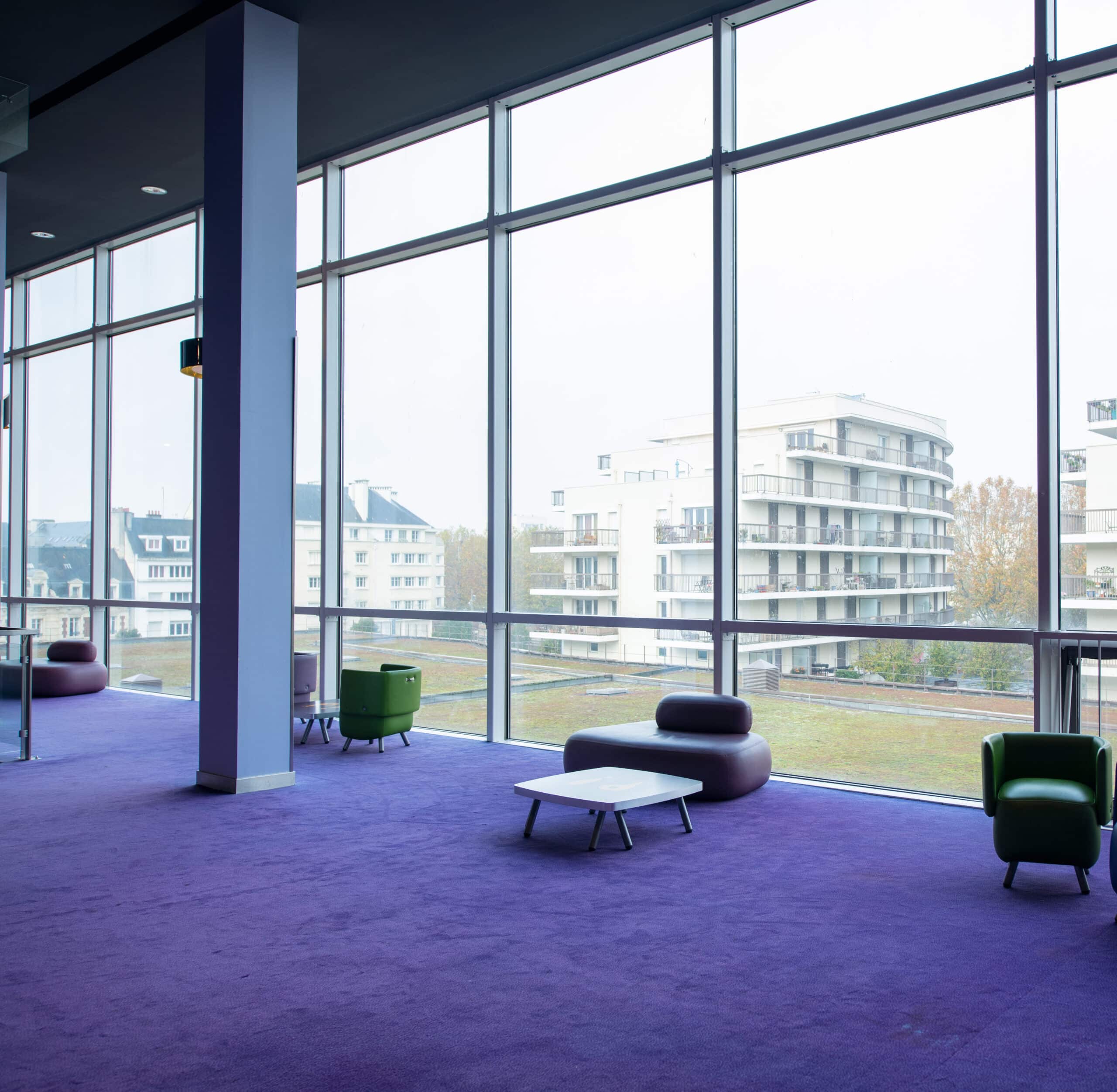 Espace lumineux avec un tapis violet, des fauteuils colorés, une grande baie vitrée donnant sur l'extérieur.