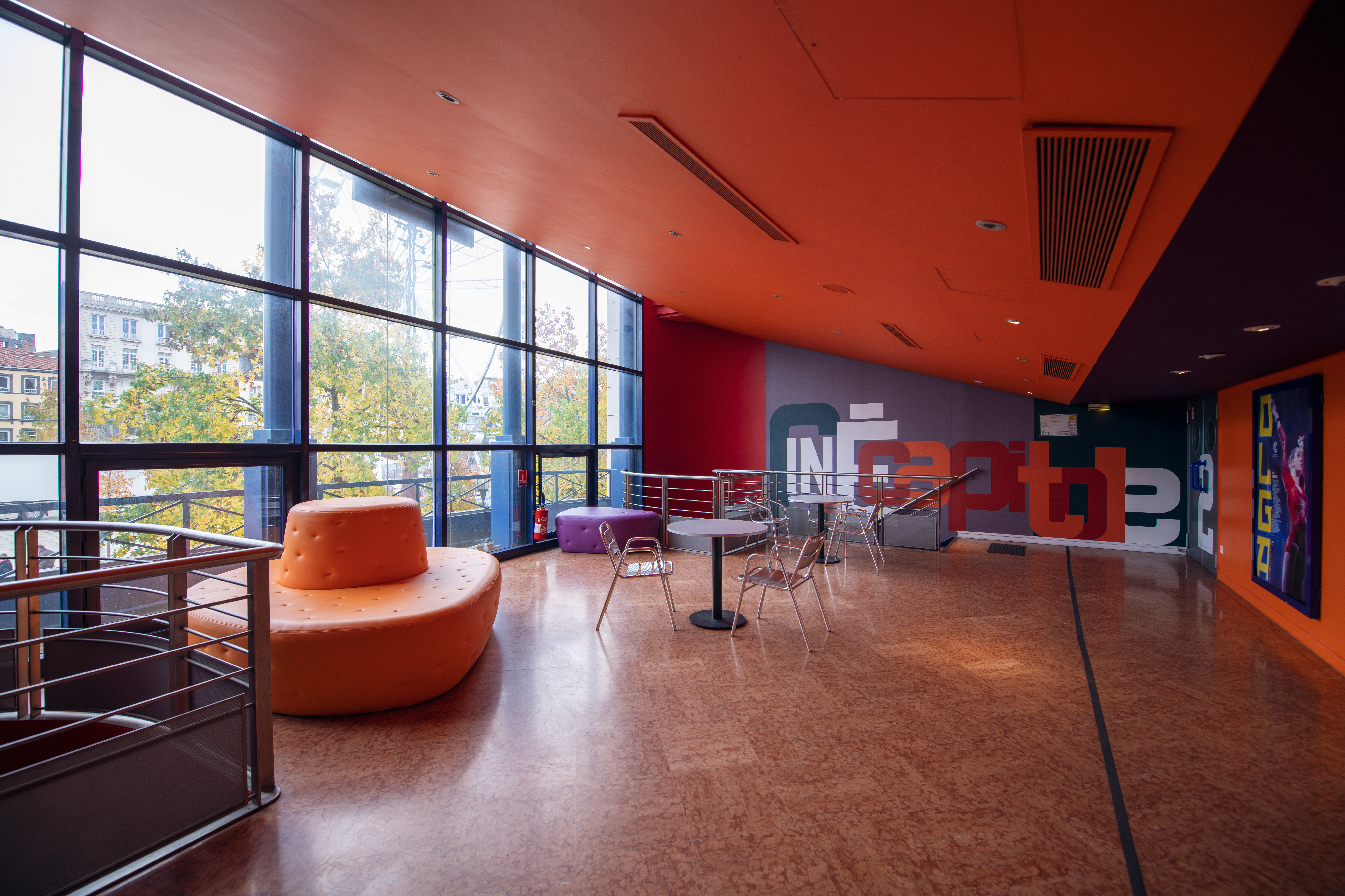 Espace lumineux du cinéma avec mobilier orange et métallique face à une grande baie vitrée offrant une vue extérieure.