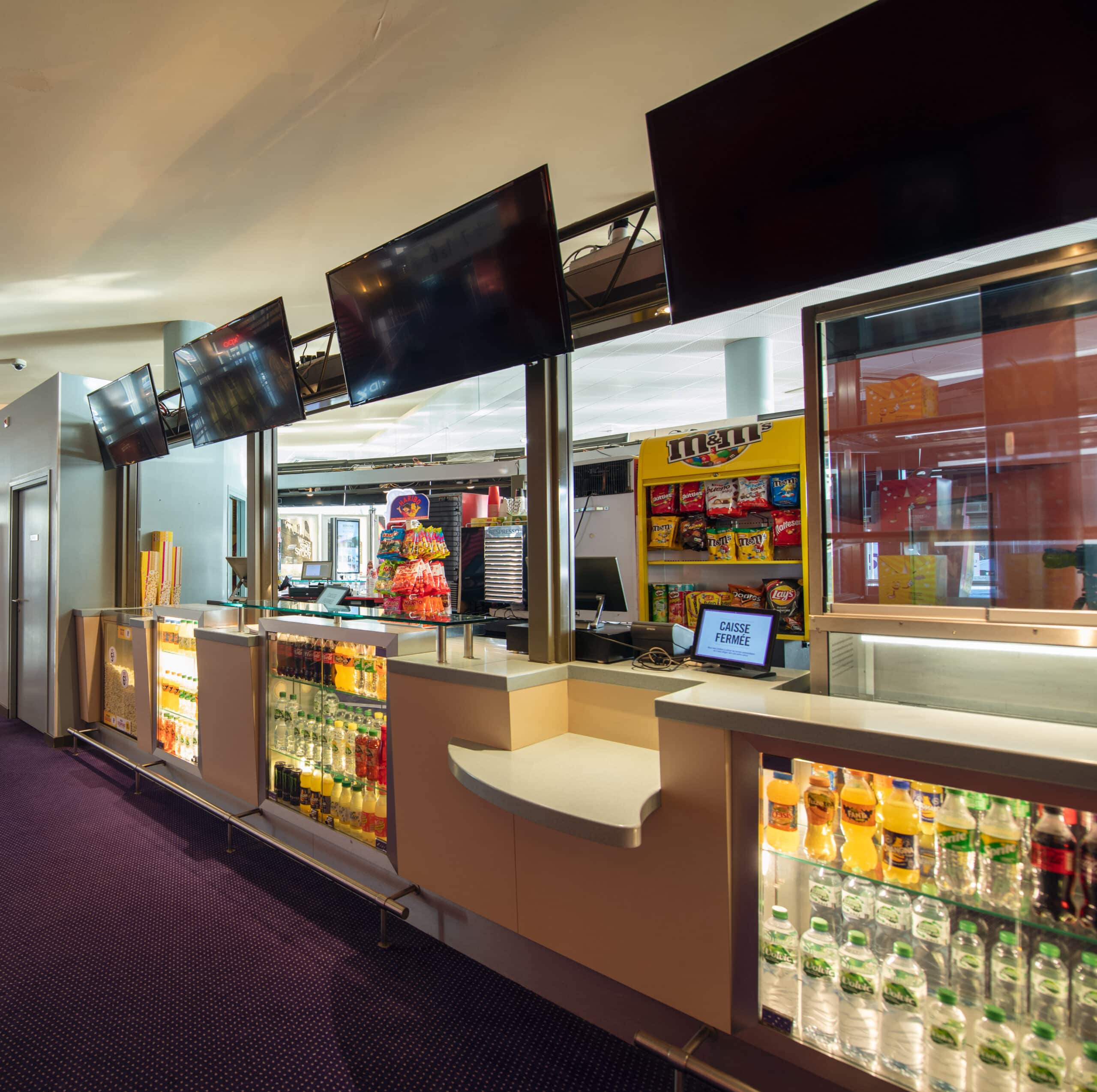 Comptoir de confiseries du cinéma avec boissons, snacks et écrans suspendus.