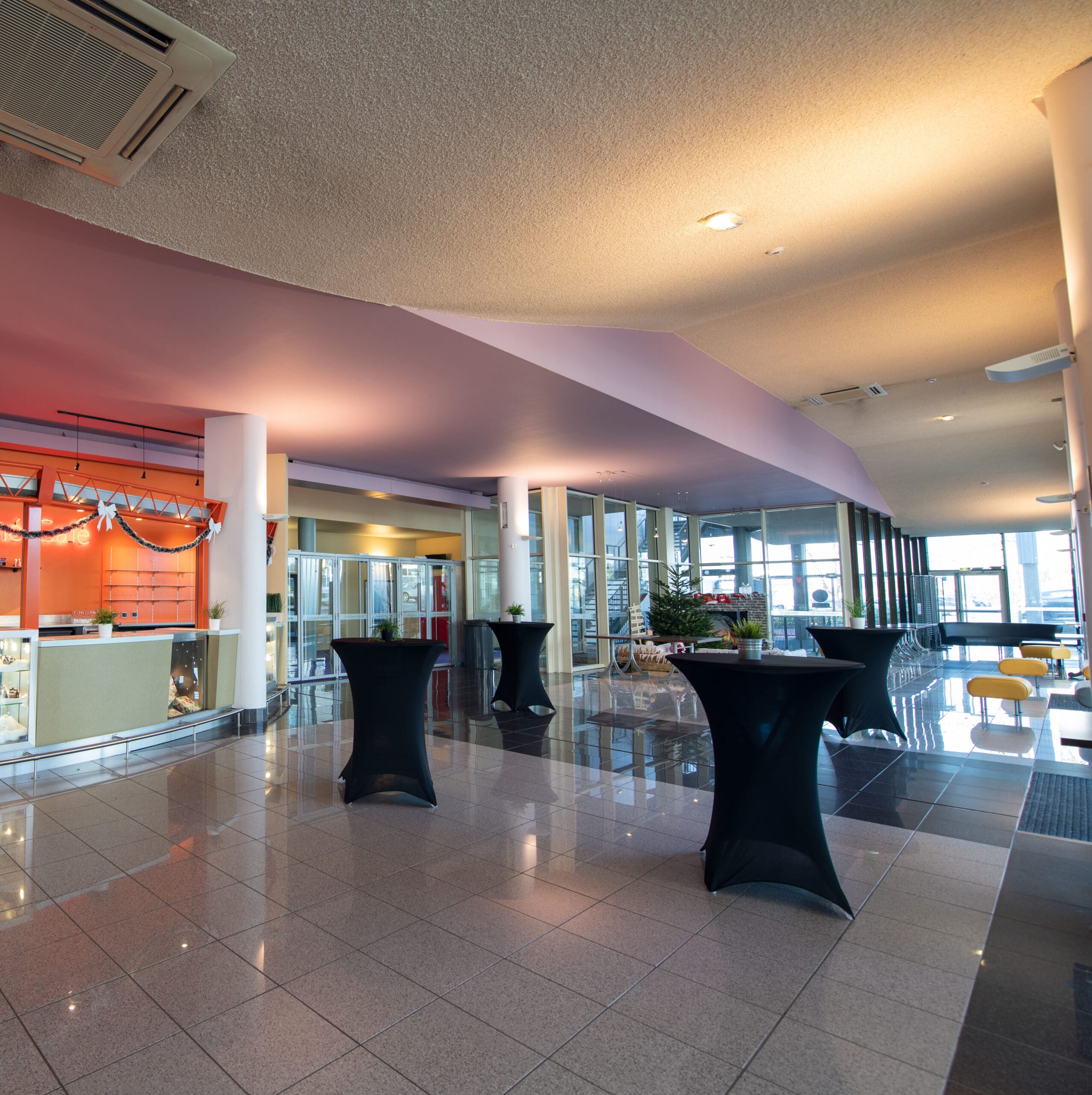 Hall spacieux avec un comptoir décoré, des tables hautes noires, une décoration festive, et des baies vitrées laissant entrer la lumière naturelle.