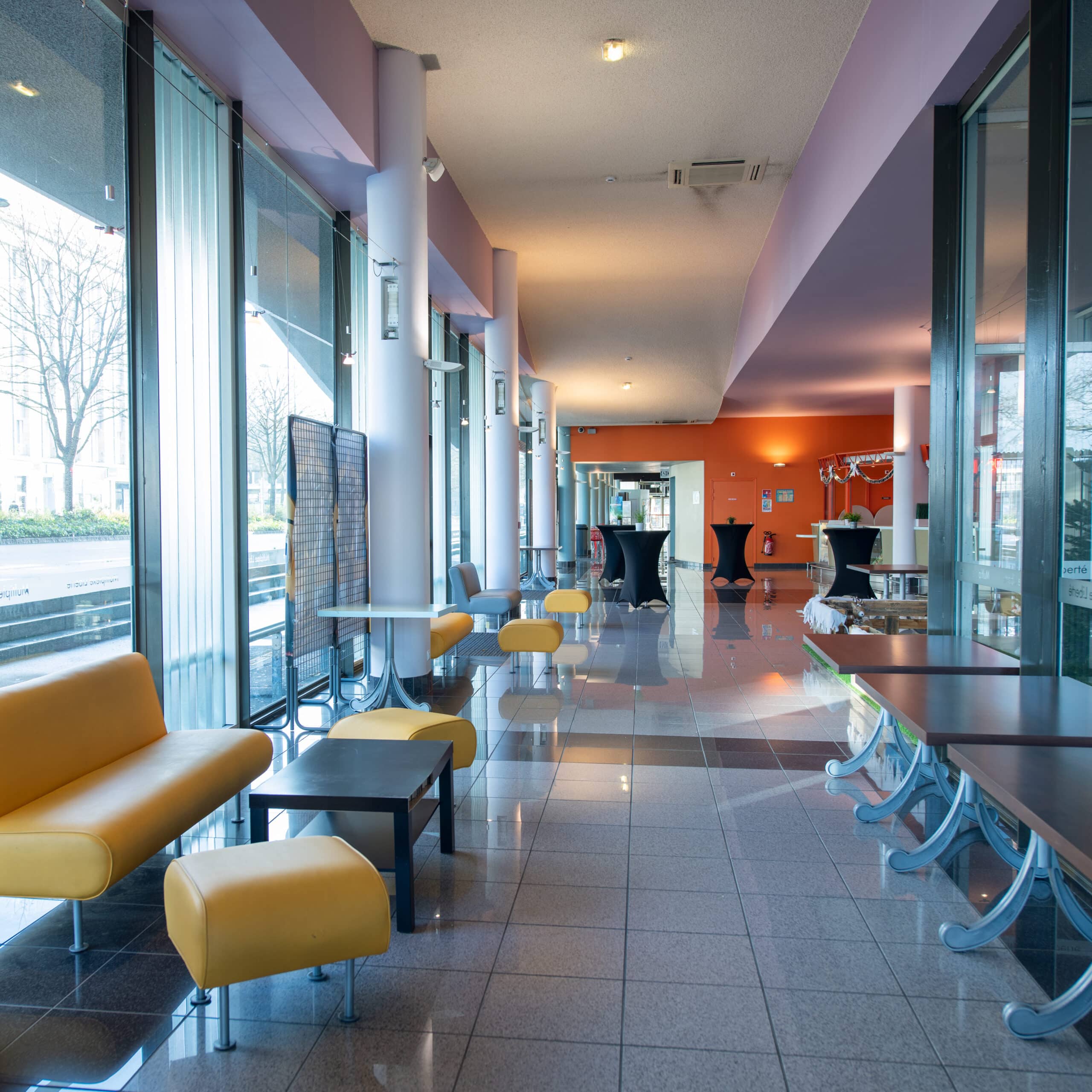 Espace lumineux avec des sièges jaunes, des petites tables, et une grande baie vitrée offrant une vue sur l'extérieur.