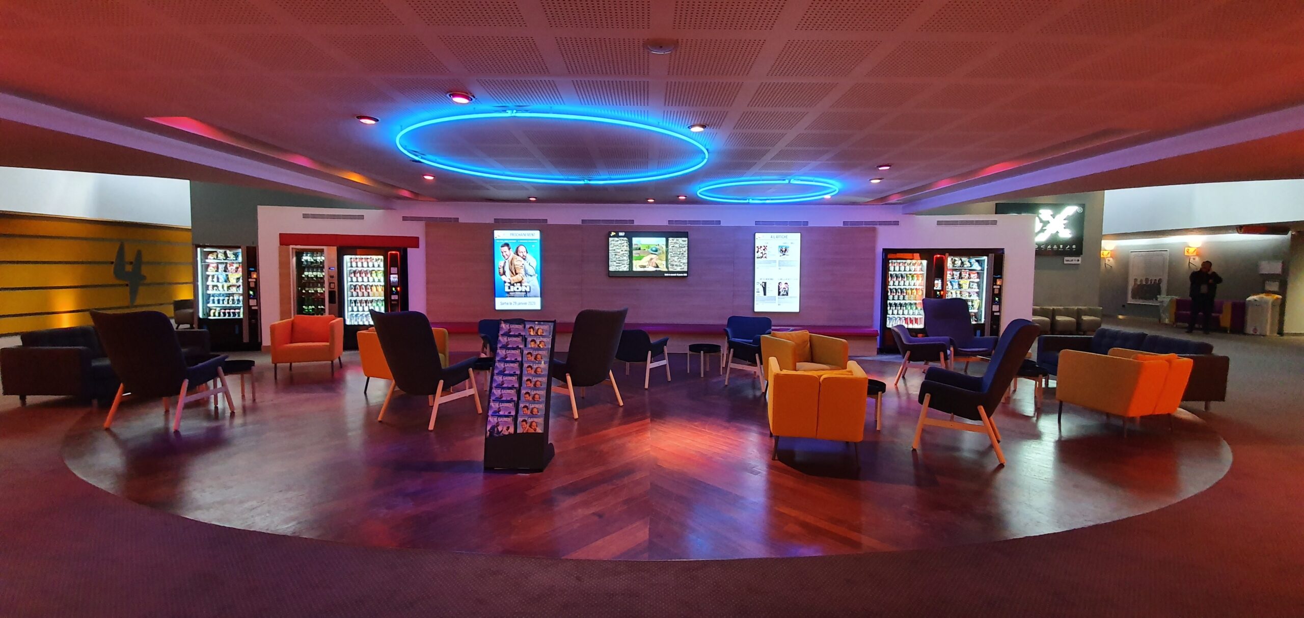 Espace de réception du cinéma avec fauteuils colorés, éclairage LED bleu et distributeurs automatiques.