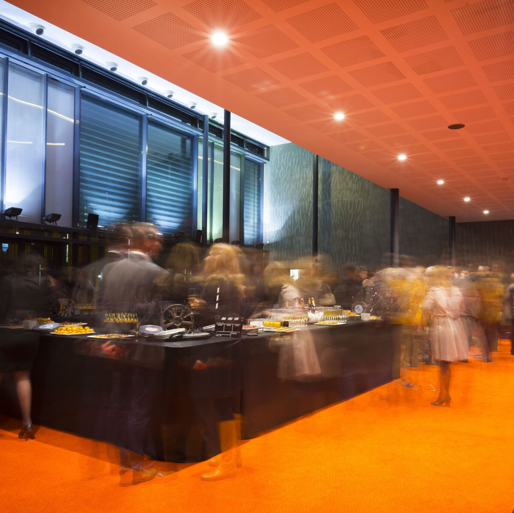 Salle de réception avec buffet élégant proposant divers plats et amuse-bouches. Les grandes baies vitrées, la moquette orange et l'éclairage offrent une ambiance lumineuse et moderne.