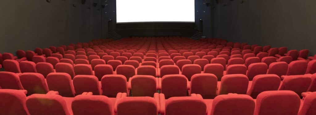 Vue depuis l’arrière d'une salle de cinéma avec ses rangées de fauteuils en velours rouges orientés vers l’écran.