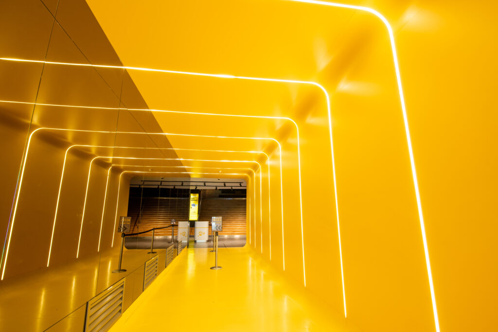 Couloir lumineux entièrement jaune avec des lignes LED intégrées aux murs et au plafond, menant à une entrée équipée de panneaux d’information.