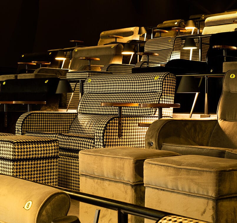 Fauteuils de cinéma haut de gamme en tissu à motifs pied-de-poule et velours, équipés de repose-pieds, tables individuelles et éclairage chaleureux.