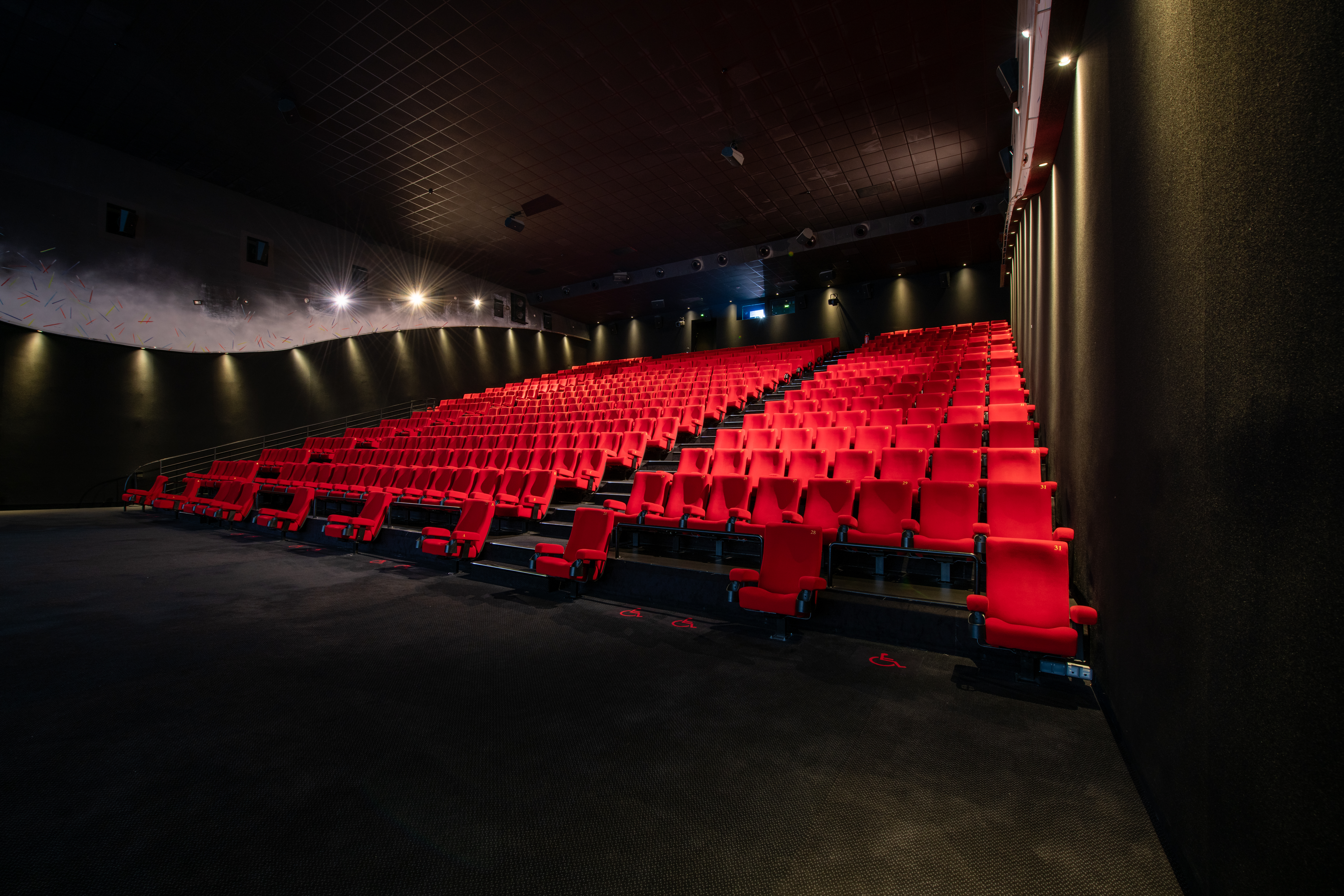 Vue latérale de la salle de cinéma montrant les fauteuils en velours rouges et l’agencement des rangées ainsi que son espace scénique.