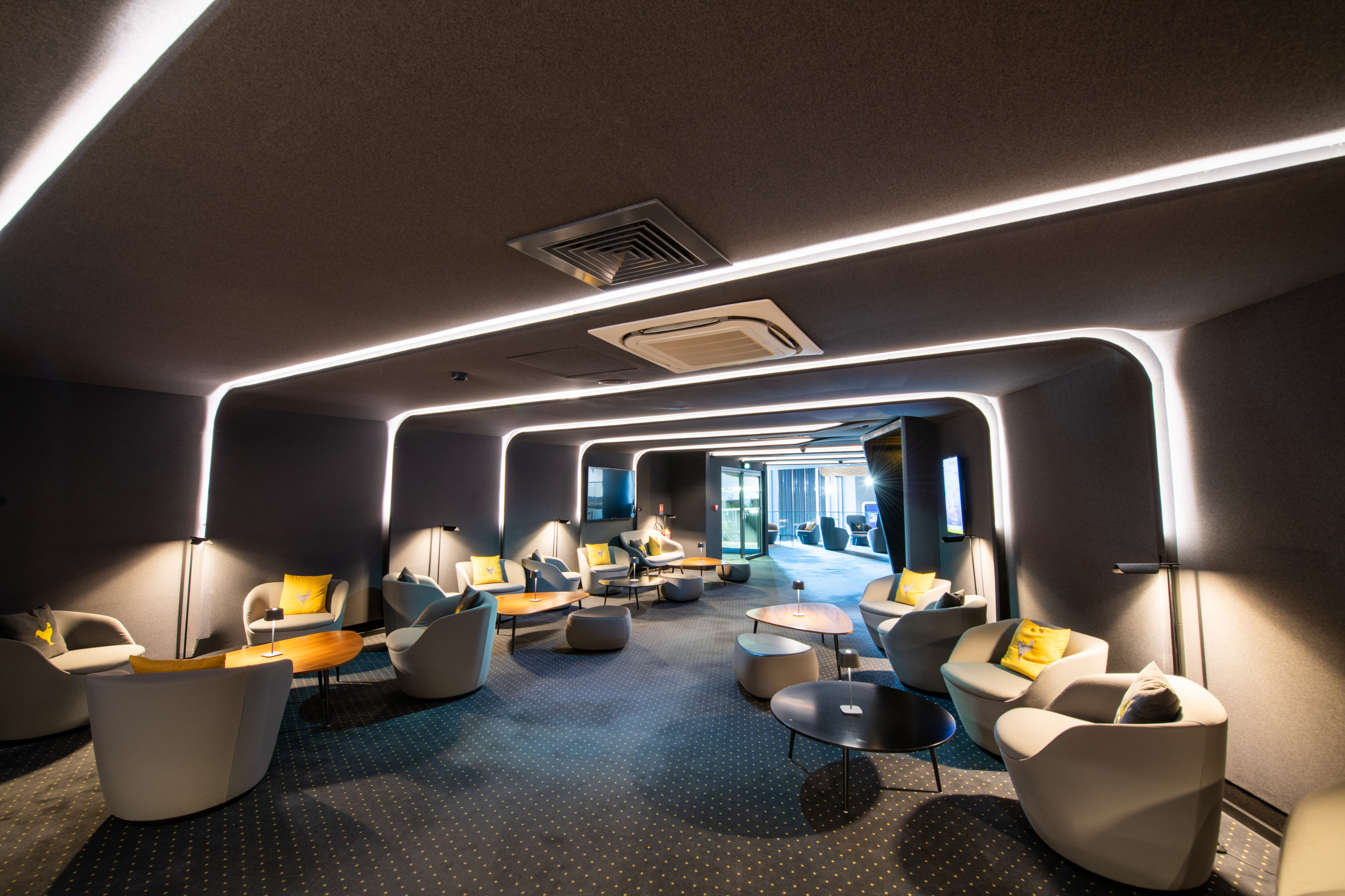 Espace cocktail du cinéma, éclairé par des néons blancs et des lampes de table à lumière chaude, avec du mobilier gris et jaune.