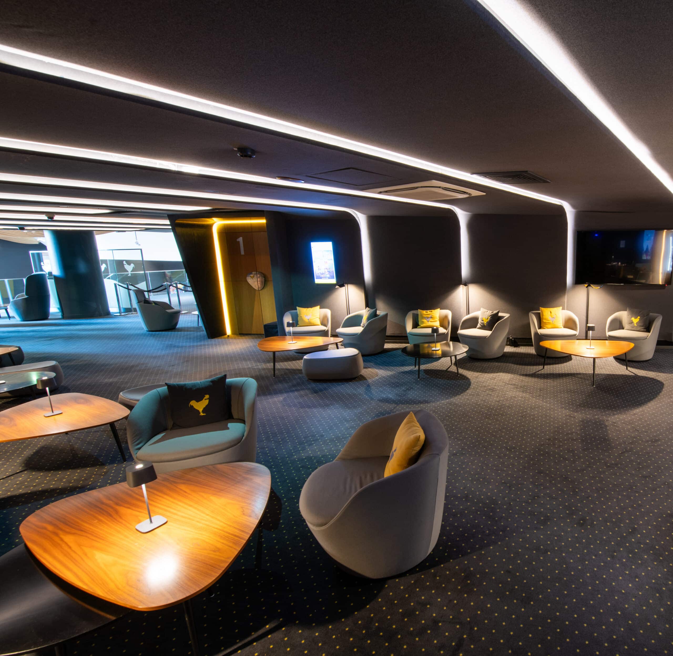 Espace cocktail du cinéma, éclairé par des néons blancs et des lampes de table à lumière chaude, avec du mobilier gris et jaune.