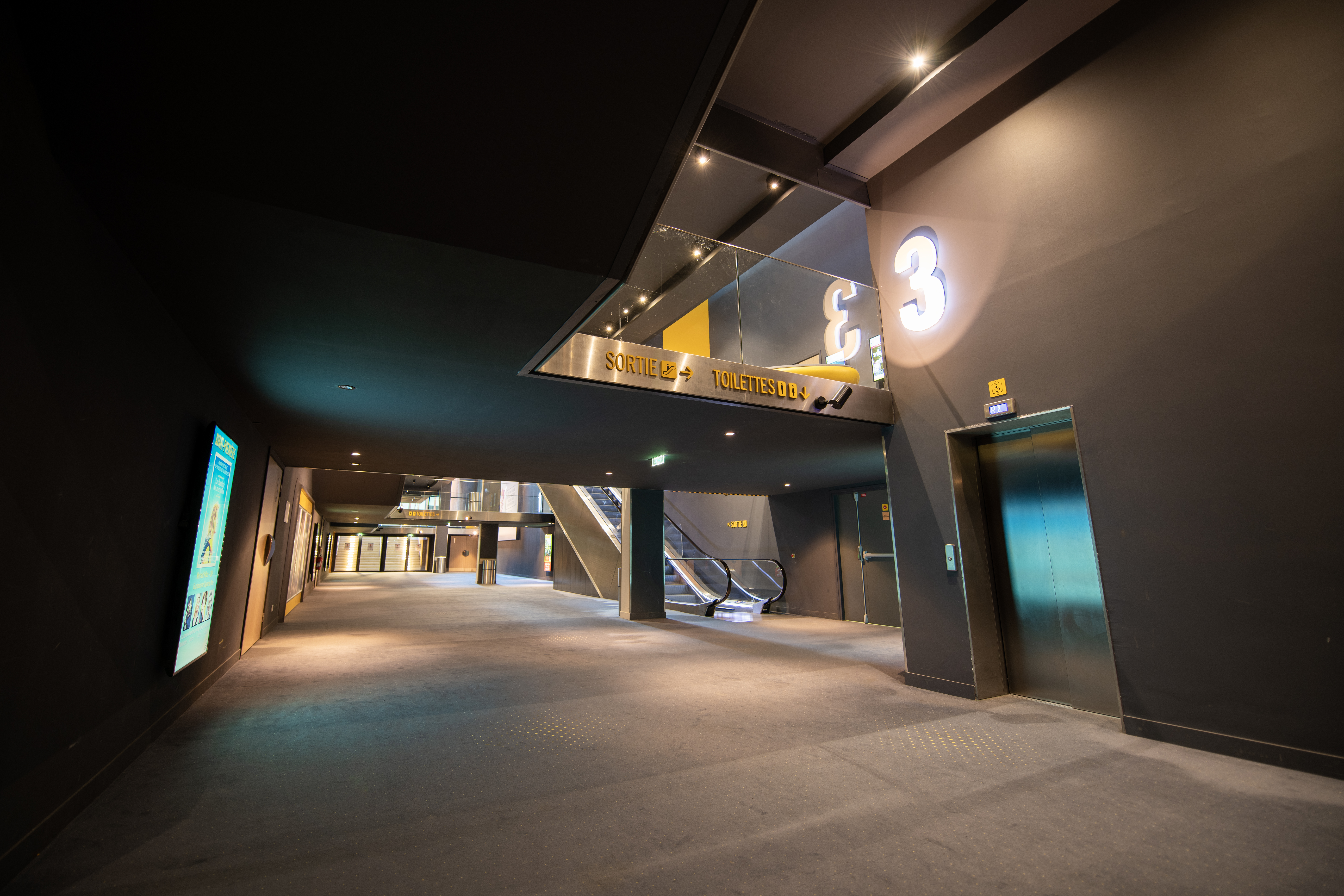 Couloir spacieux dans un cinéma avec des panneaux d'indication pour la sortie et les toilettes, un escalator, un ascenseur et des affiches de films éclairées.