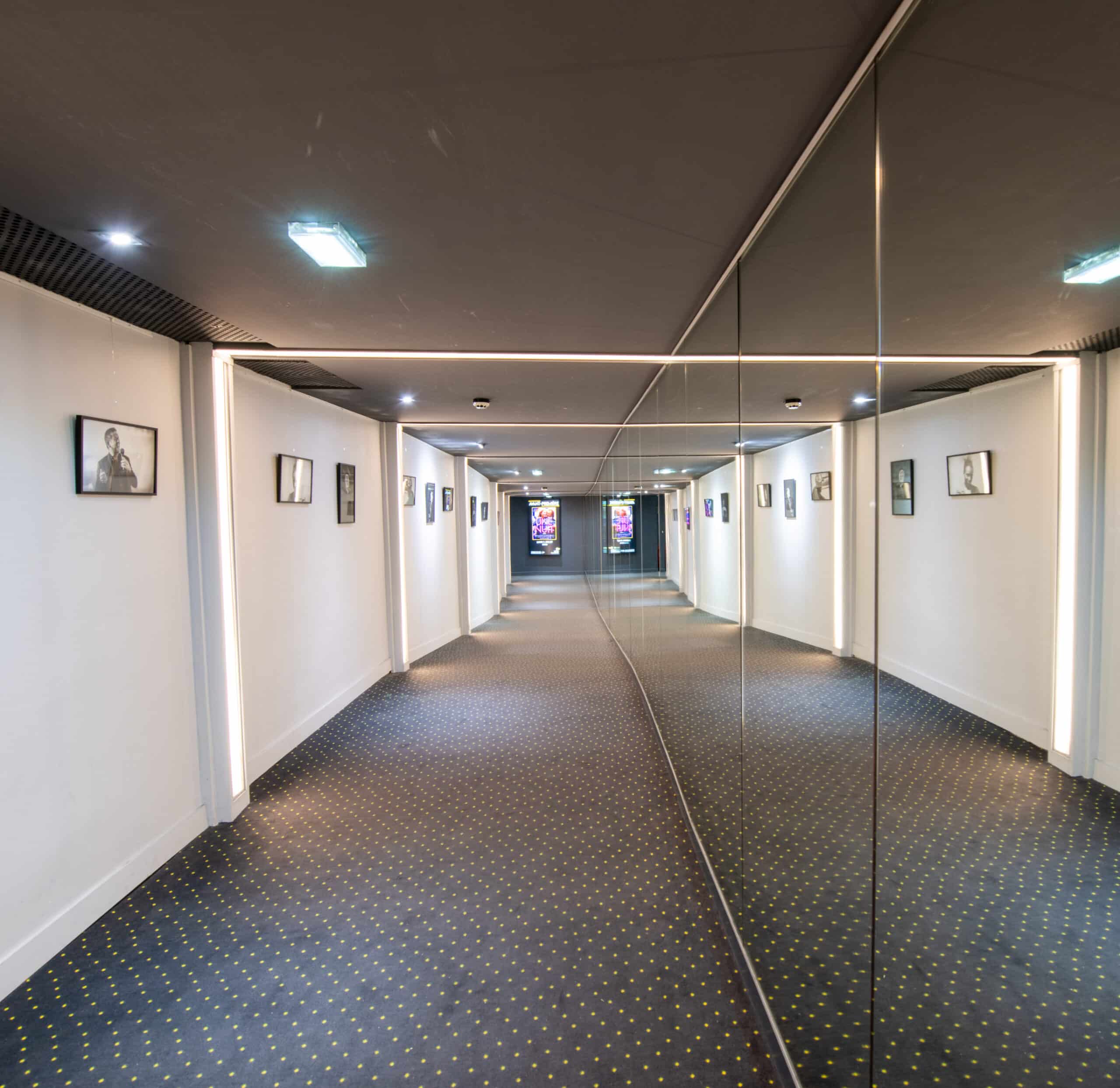 Couloir lumineux avec moquette à motifs, murs décorés de cadres et miroir sur toute la longueur.