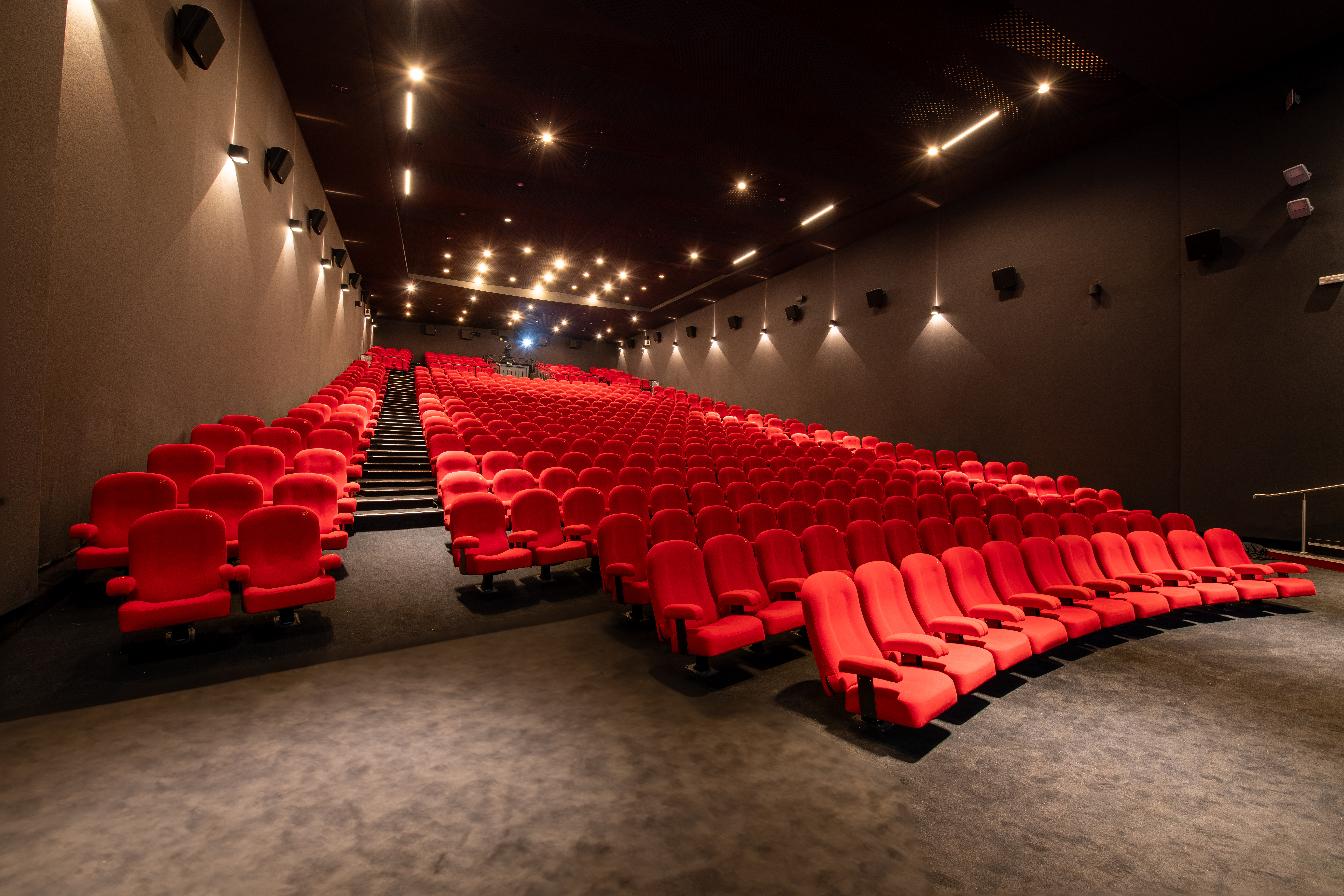 Vue latérale de la salle de cinéma montrant les fauteuils en velours rouges et l’agencement des rangées ainsi que son espace scénique.