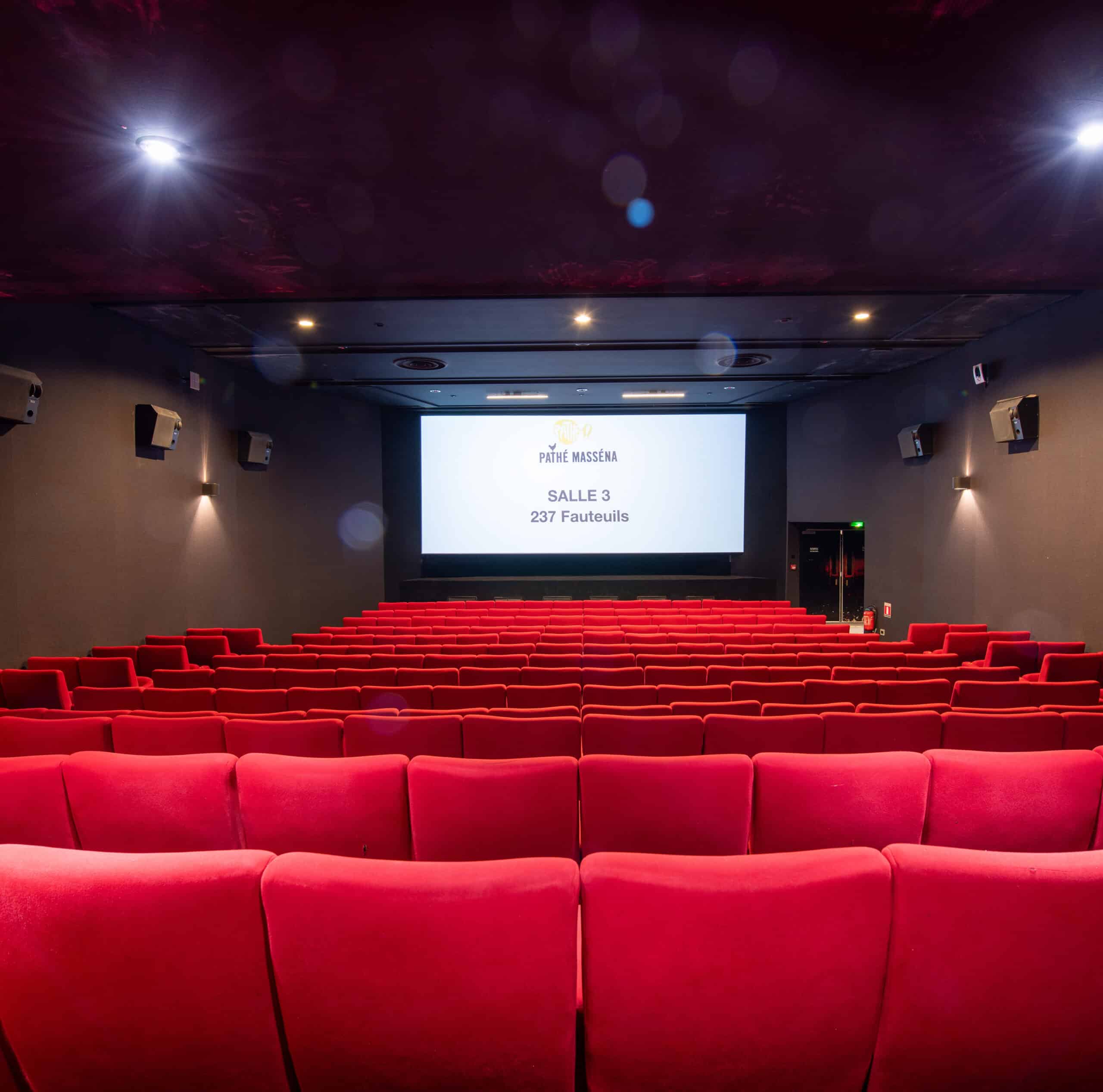 Vue depuis l’arrière de la salle 5 du cinéma Pathé Masséna avec rangées de fauteuils en velours rouges orientés vers l’écran.