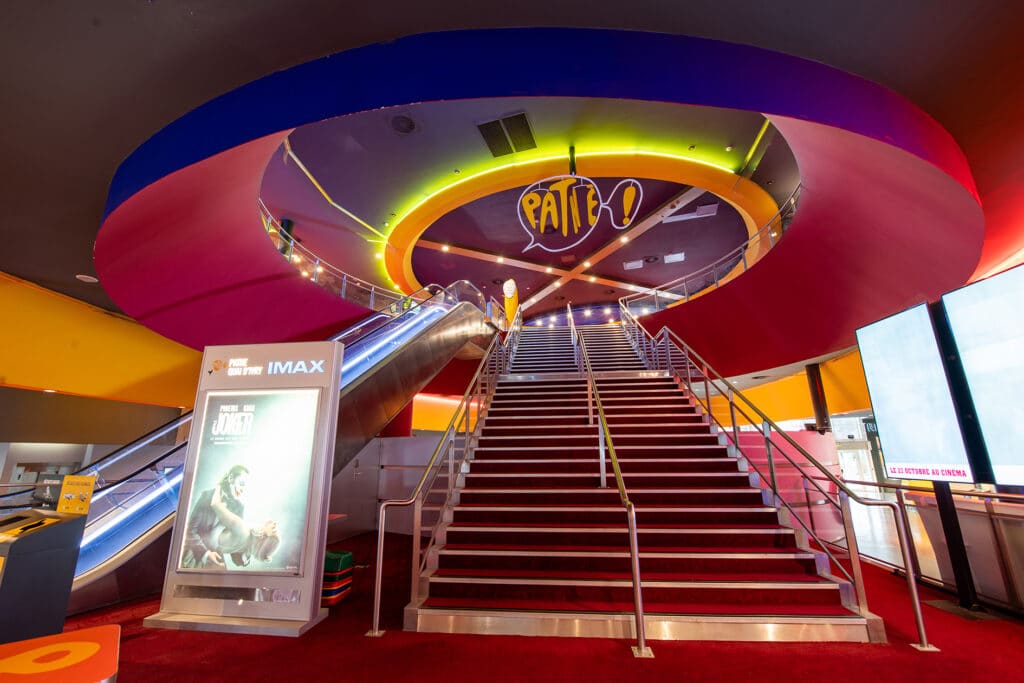 Escalier majestueux bordé de moquette rouge, accompagné d’un escalator, enseigne Pathé lumineuse suspendue et affichage IMAX à proximité