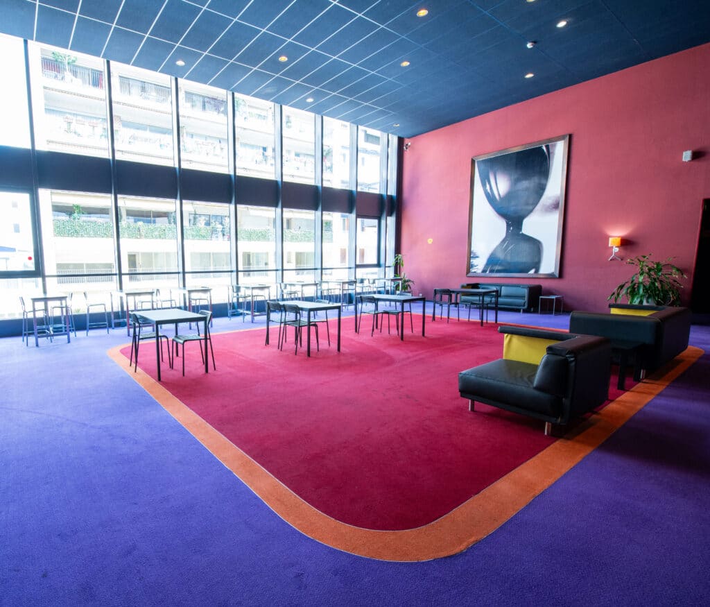 Espace cocktail du cinéma à la lumière du jour grâce à ses grandes baies vitrées, équipé d'un mobilier de réception et de salon.