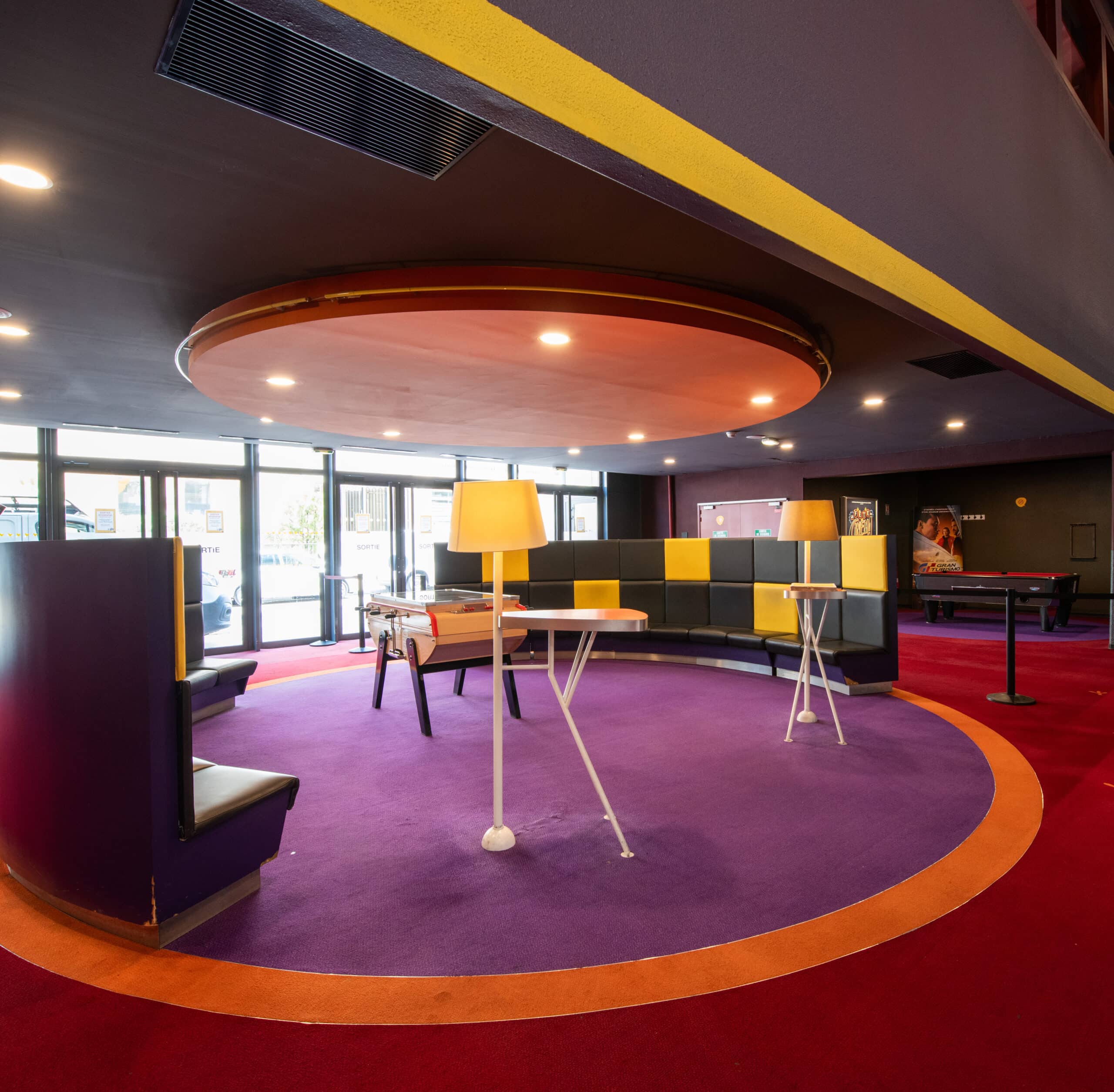 Espace intérieur coloré avec un sol en moquette violet entouré d’un cercle orange. Des banquettes arrondies noires et jaunes occupent le centre, accompagnées de petites tables et lampes modernes. Le plafond sombre est muni d’un élément circulaire rétroéclairé. De larges fenêtres apportent de la lumière naturelle.