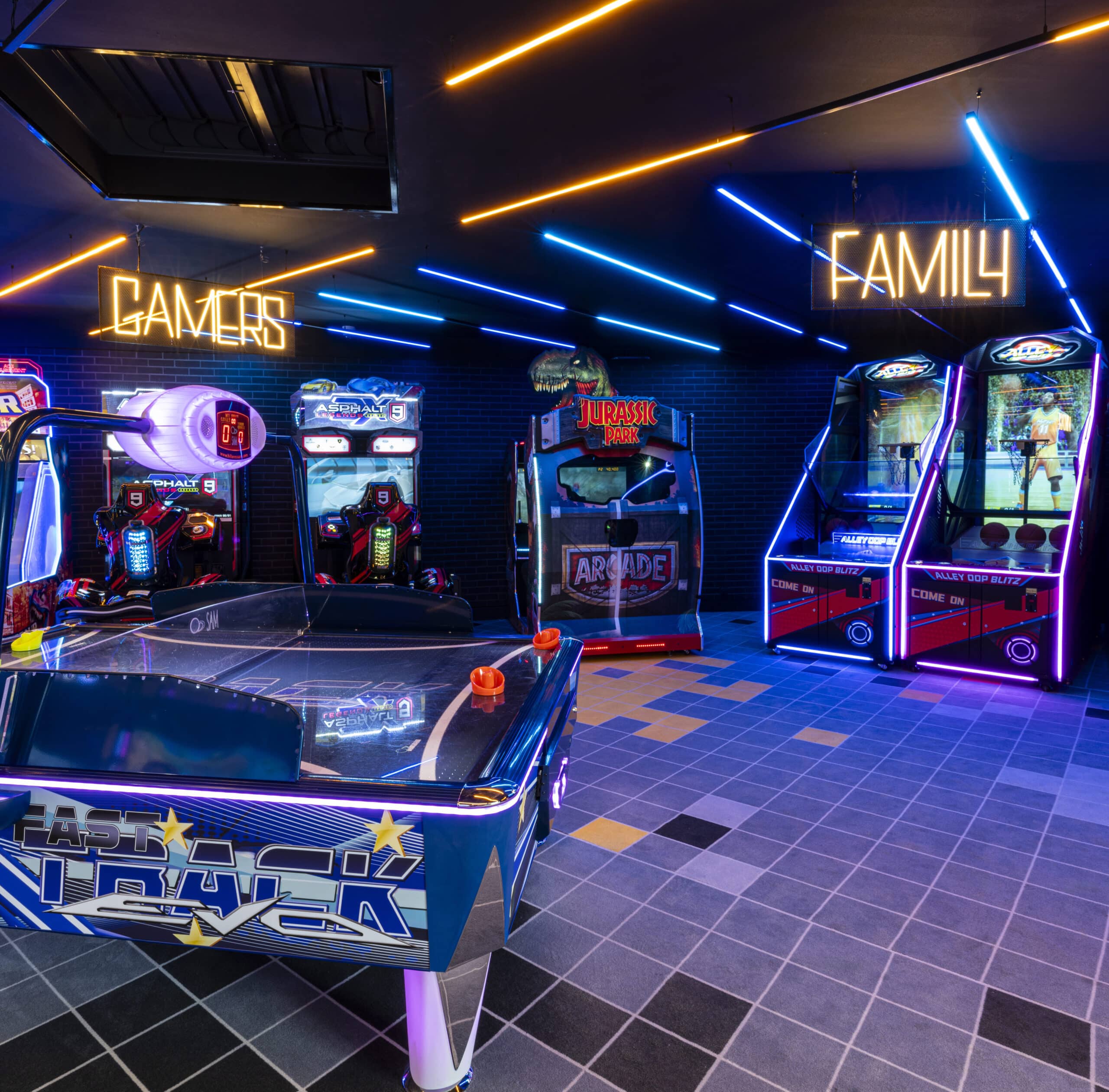 Salle Pathé Games aux lumières néon bleues et jaunes, avec plusieurs machines d’arcade colorées, dont un jeu de hockey sur air en premier plan, des bornes de course Asphalt 9, une machine Jurassic Park, et deux jeux de basket-ball Alley Oop Blitz, séparées en zones ‘Gamers’ et ‘Family’. »« Espace Pathé Games avec différentes machines d’arcade et de jeux vidéo, éclairage néon coloré, ambiance moderne et ludique, proposant des jeux pour tous les âges dans un cadre convivial.