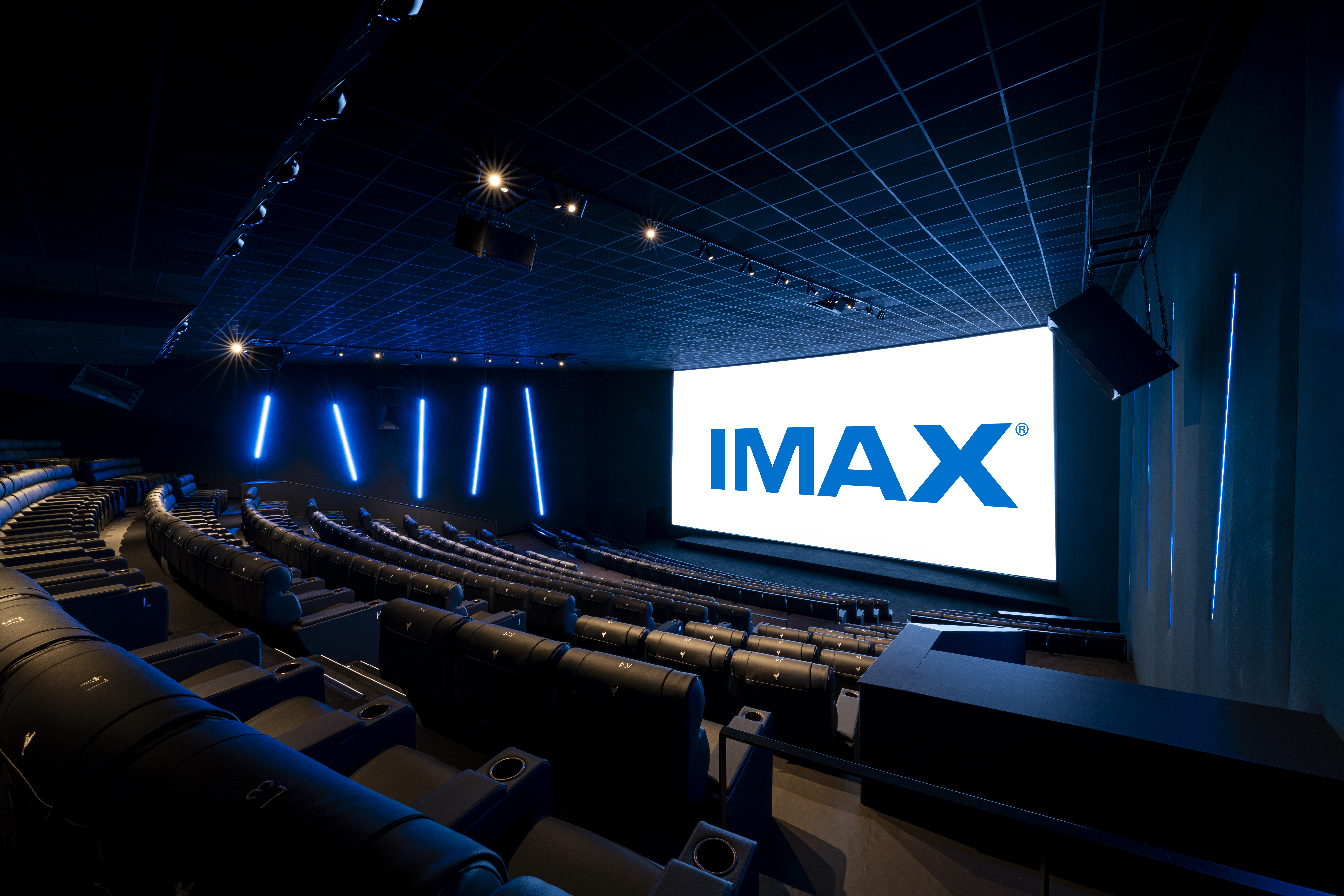 Vue de derrière d'une salle de cinéma équipée de la technologie IMAX montrant les fauteuils en cuirs noirs et l’agencement des rangées ainsi que son espace scénique.