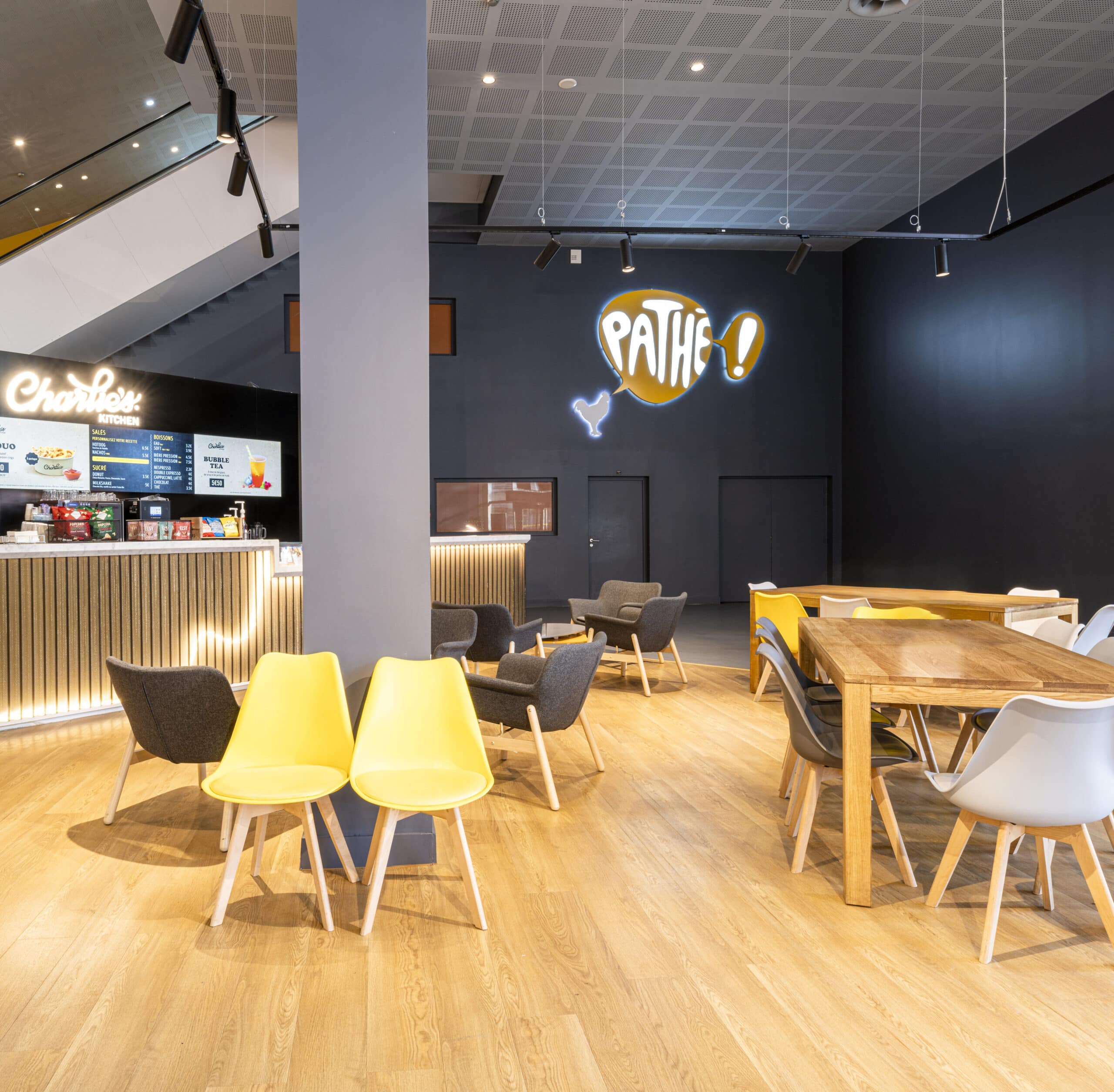 Espace accueillant dans un cinéma Pathé avec des tables en bois entourées de chaises colorées, un bar lumineux, et des étagères de confiseries à l'arrière-plan.