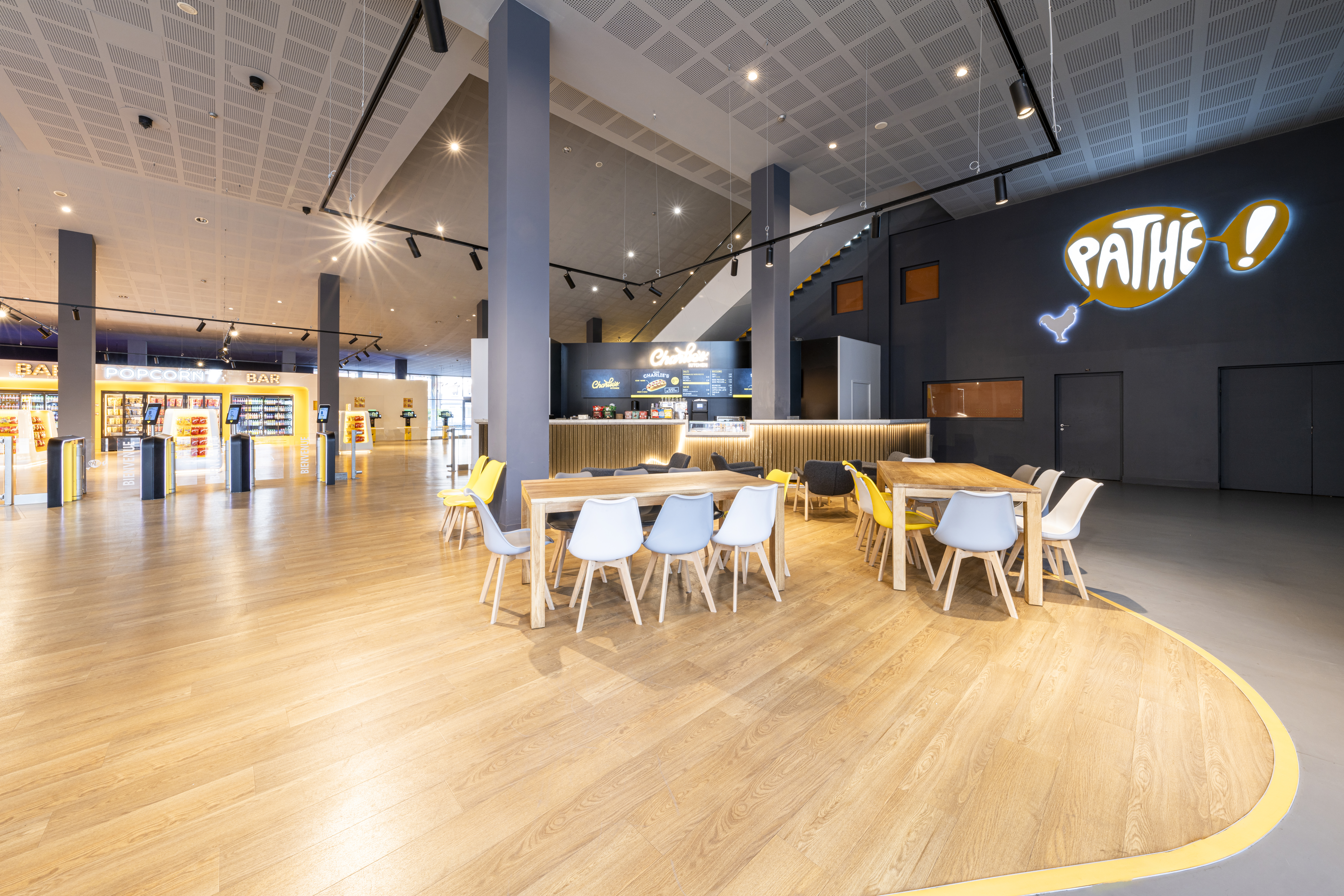 Espace accueillant dans un cinéma Pathé avec des tables en bois entourées de chaises colorées, un bar lumineux, et des étagères de confiseries à l'arrière-plan.