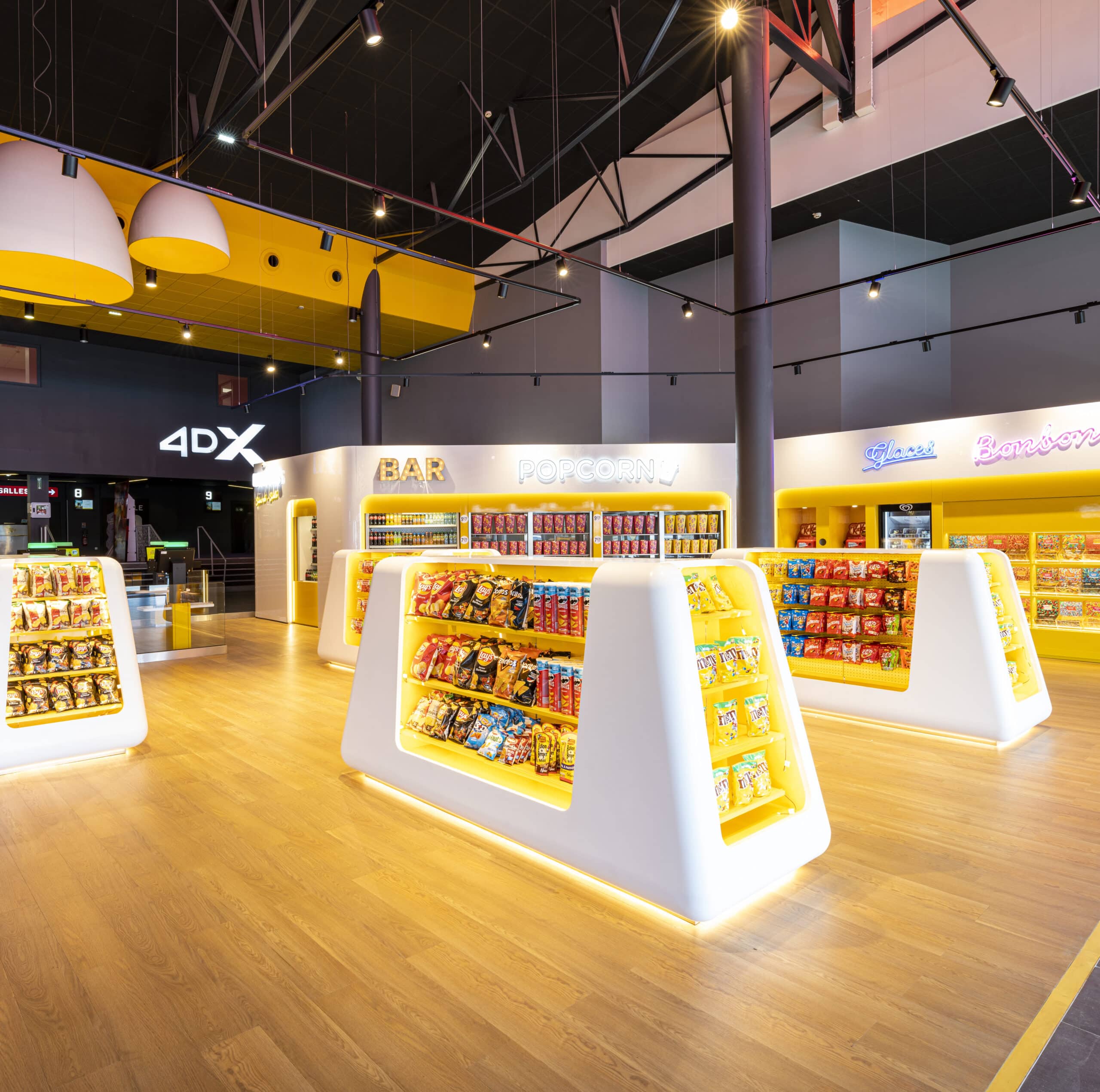 Espace lumineux dédié aux snacks avec présentoirs modernes éclairés, proposant popcorn, bonbons et autres collations, sur un sol en bois clair