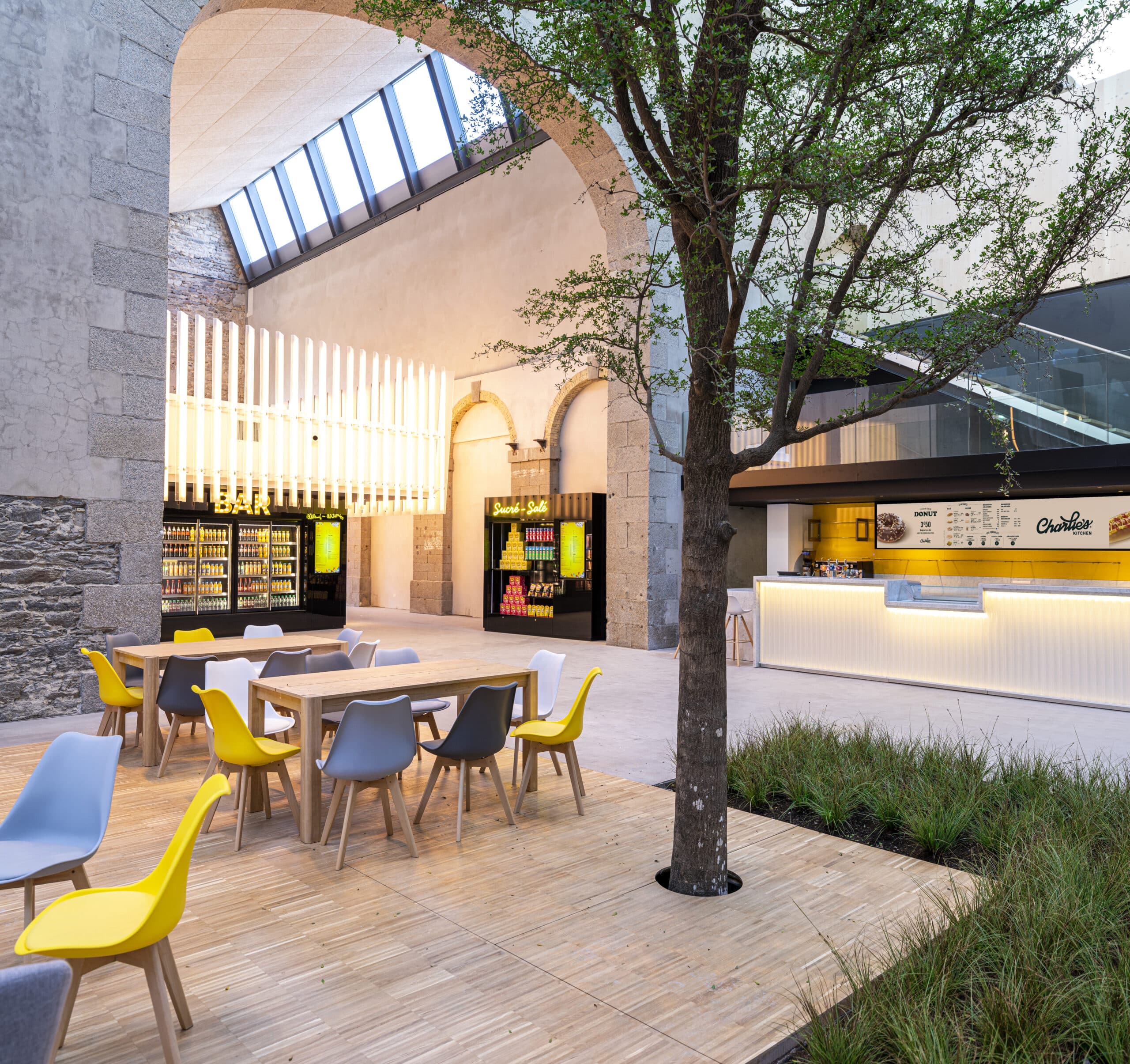 Espace extérieur convivial avec des tables et sièges colorés, un arbre central, un comptoir de restauration lumineusement décoré, et un bâtiment en bois moderne.