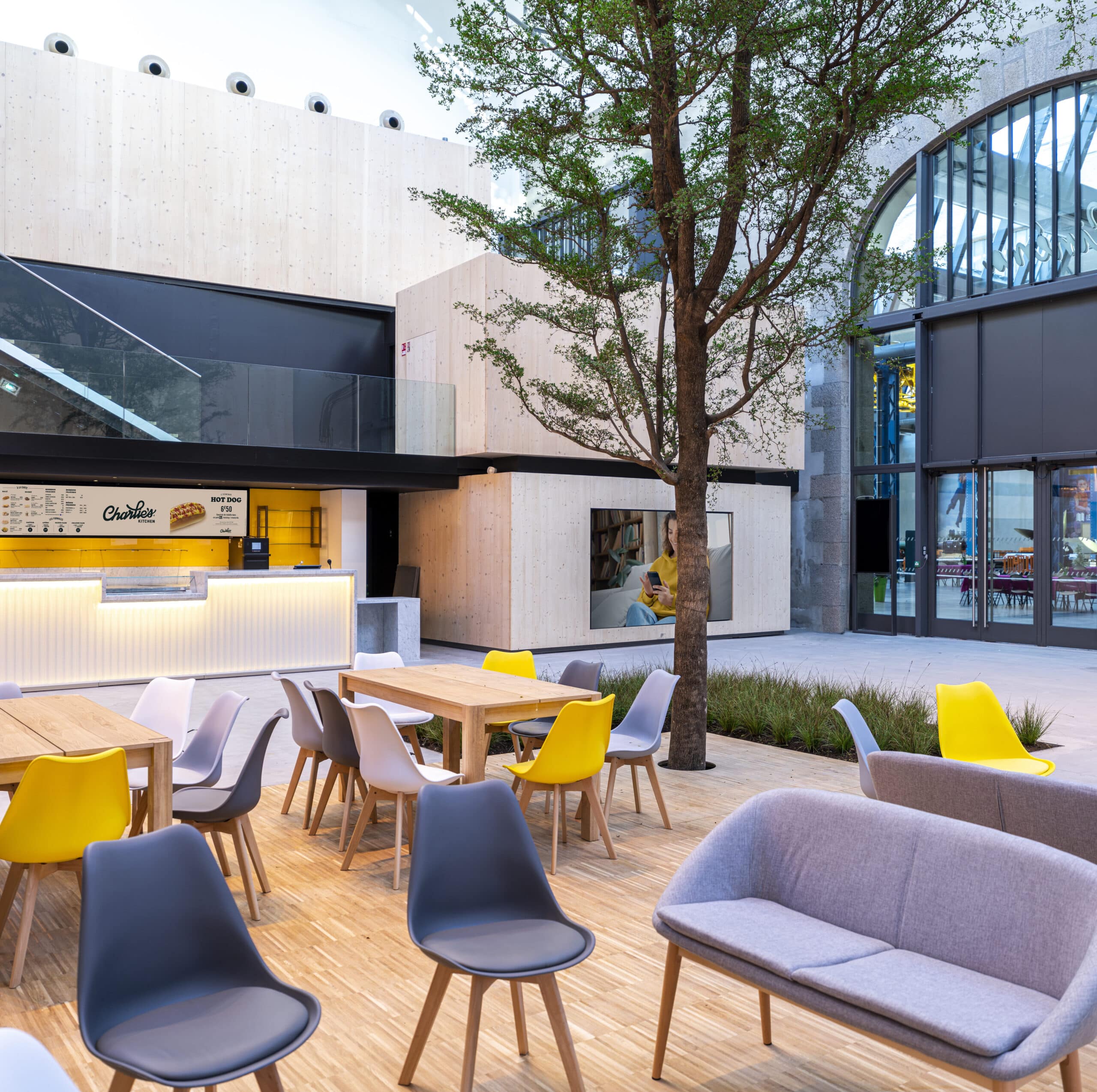 Espace extérieur convivial avec des tables et sièges colorés, un arbre central, un comptoir de restauration lumineusement décoré, et un bâtiment en bois moderne.