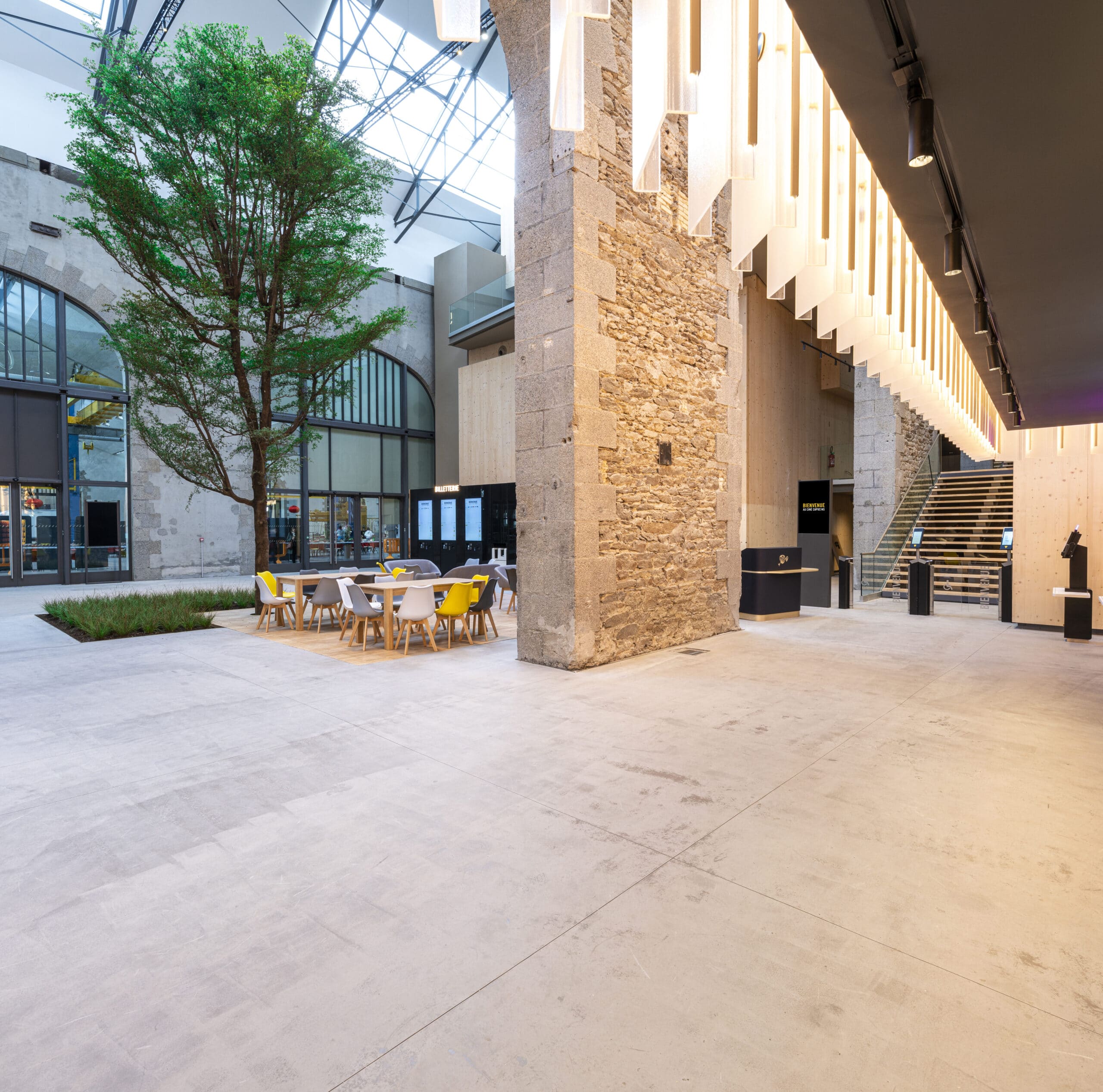 Espace ouvert avec un arbre, des tables et chaises colorées, des murs en pierre, des éclairages suspendus modernes, et un escalier en bois menant à l'étage.