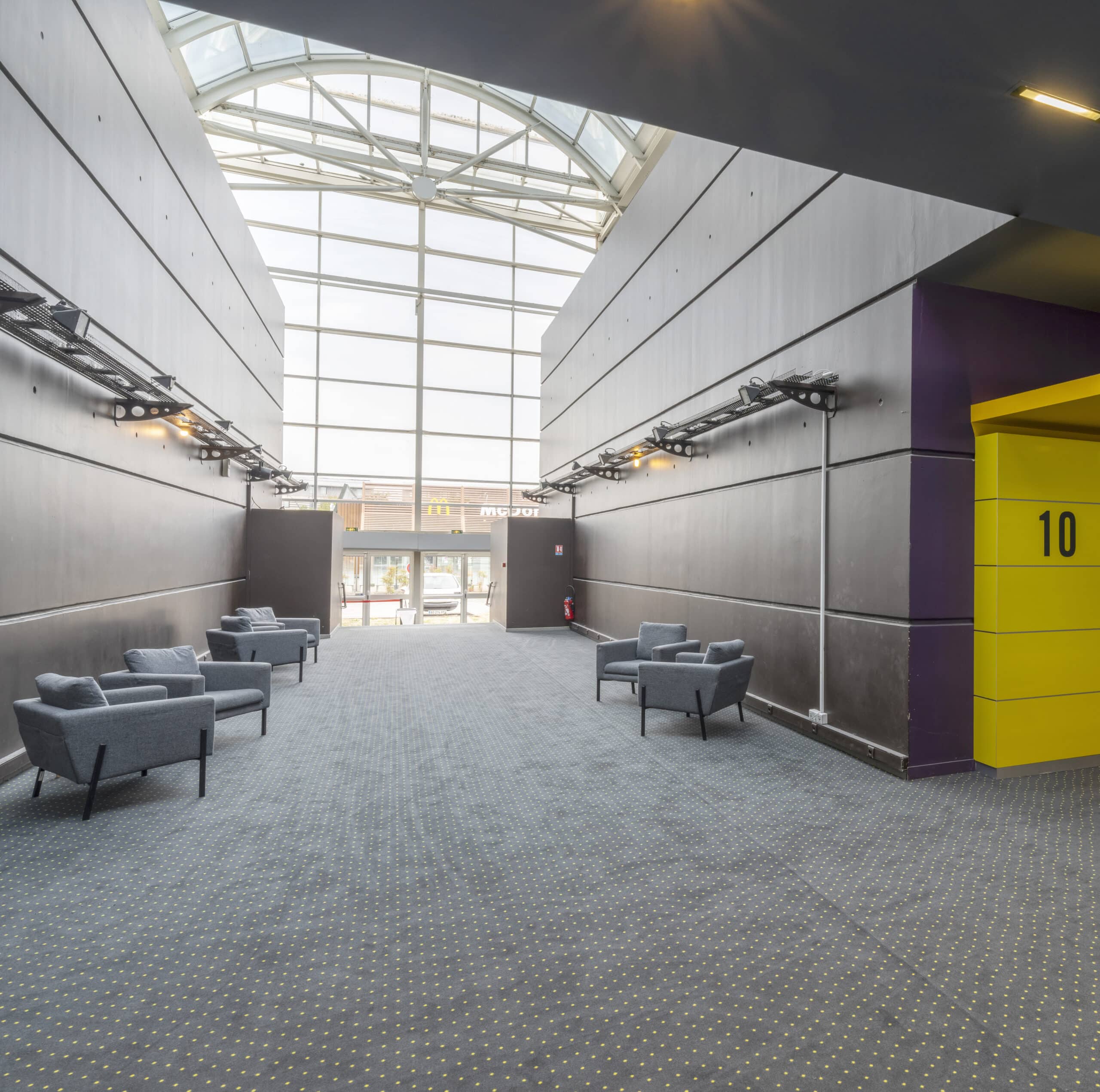 Couloir du cinéma avec salle numéro 10, fauteuils gris et grandes baies vitrées illuminant l’espace de réception.