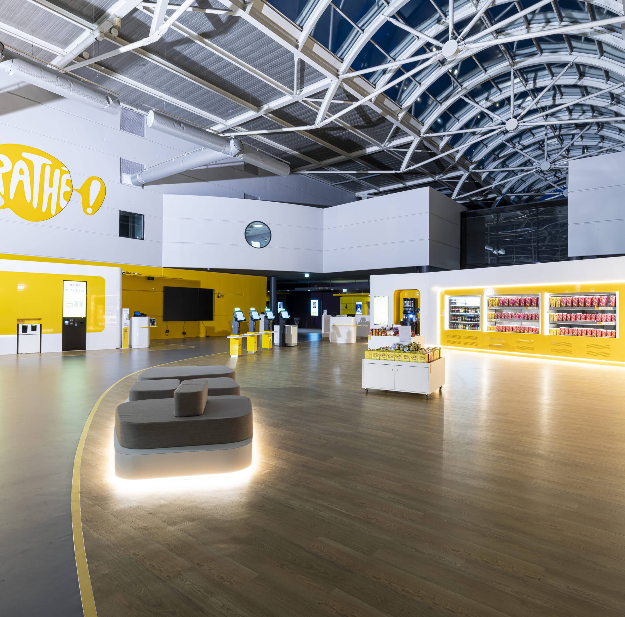 Hall du cinéma Pathé avec bornes automatiques, étalages de snacks et design lumineux sous une verrière.