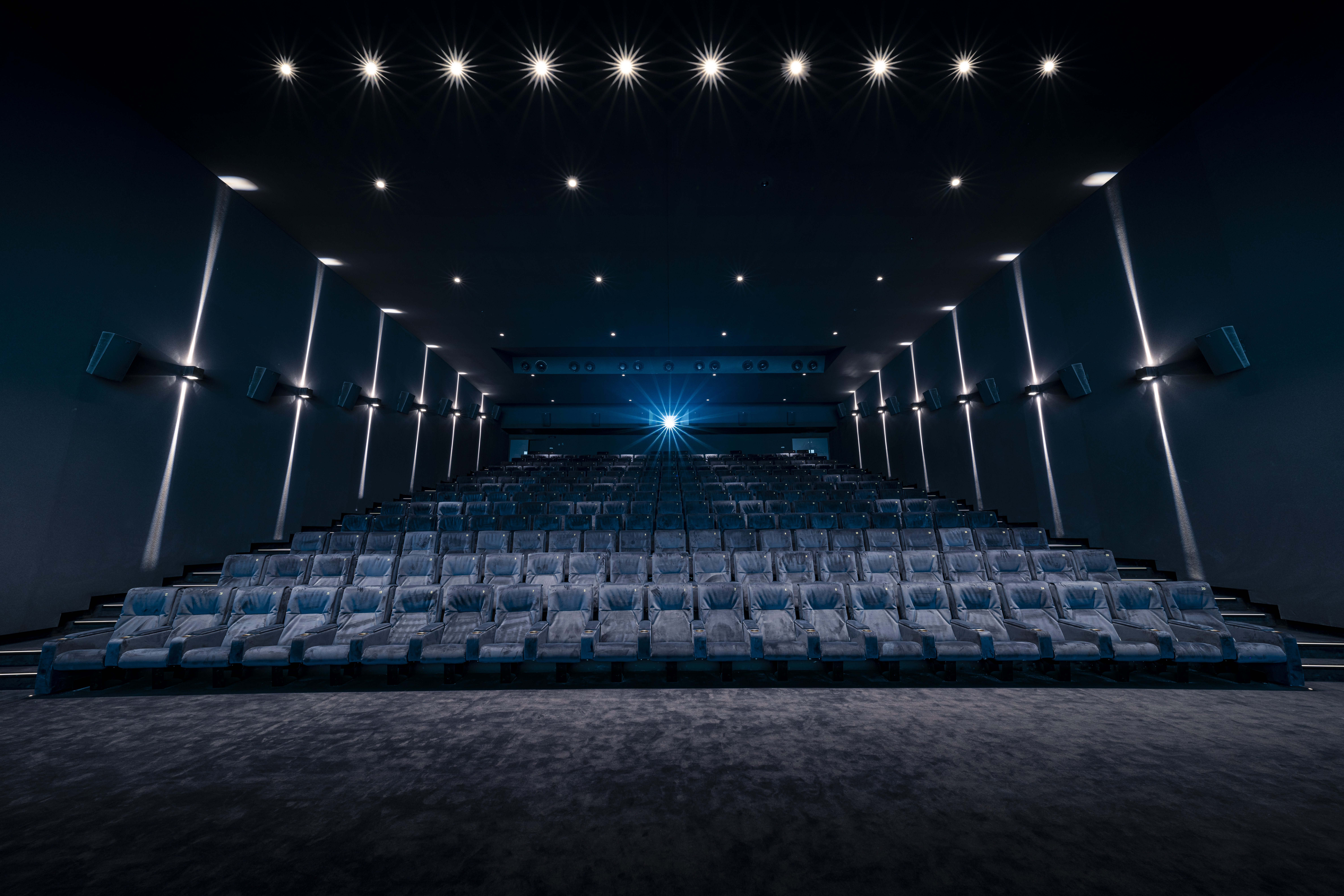 Salle de cinéma sombre et moderne avec rangées de sièges en cuir, éclairage discret sur les murs et projecteur allumé au fond, créant une ambiance immersive.