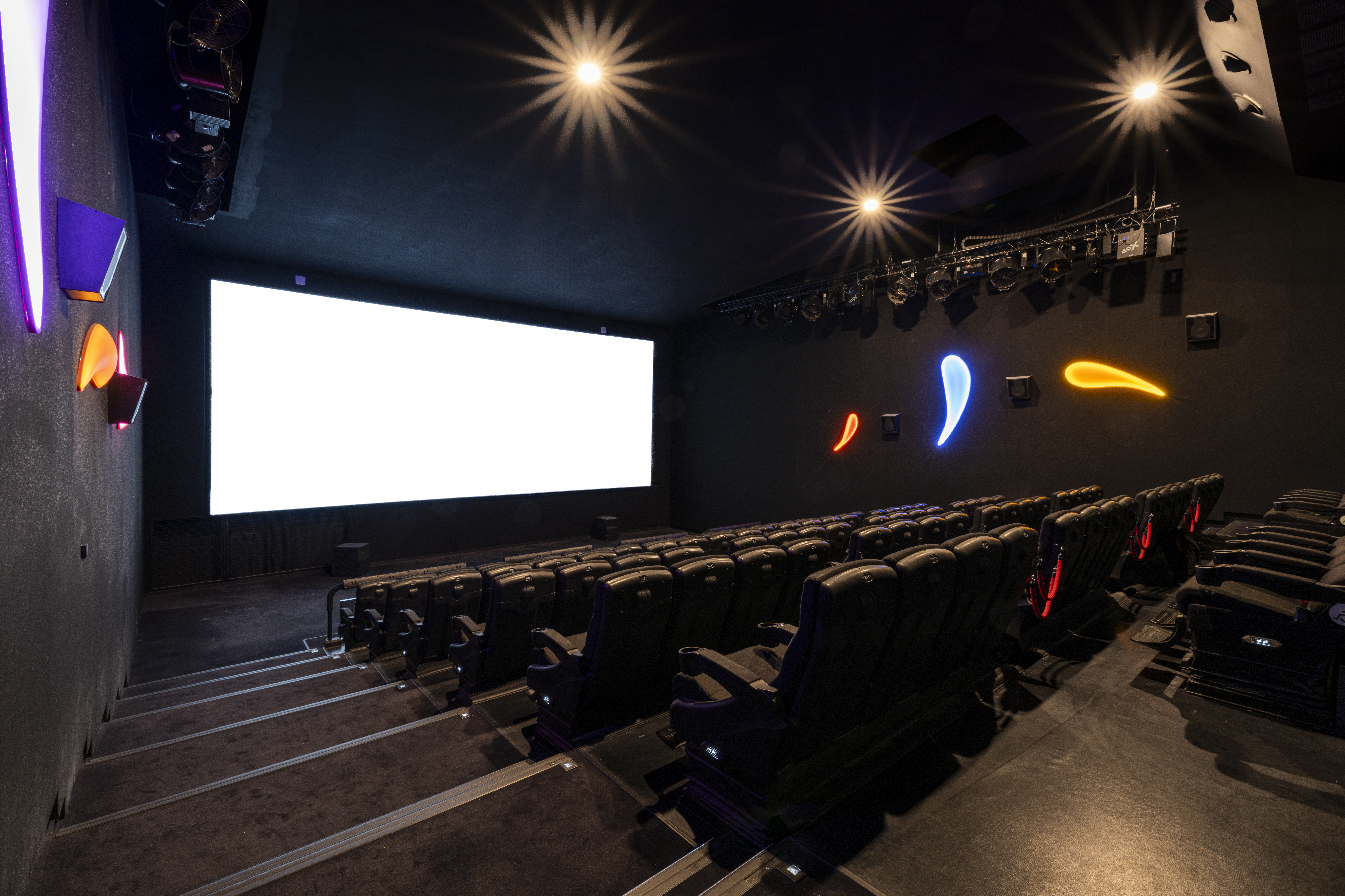 Salle de cinéma immersive avec sièges noirs, éclairages colorés sur les murs et écran géant.