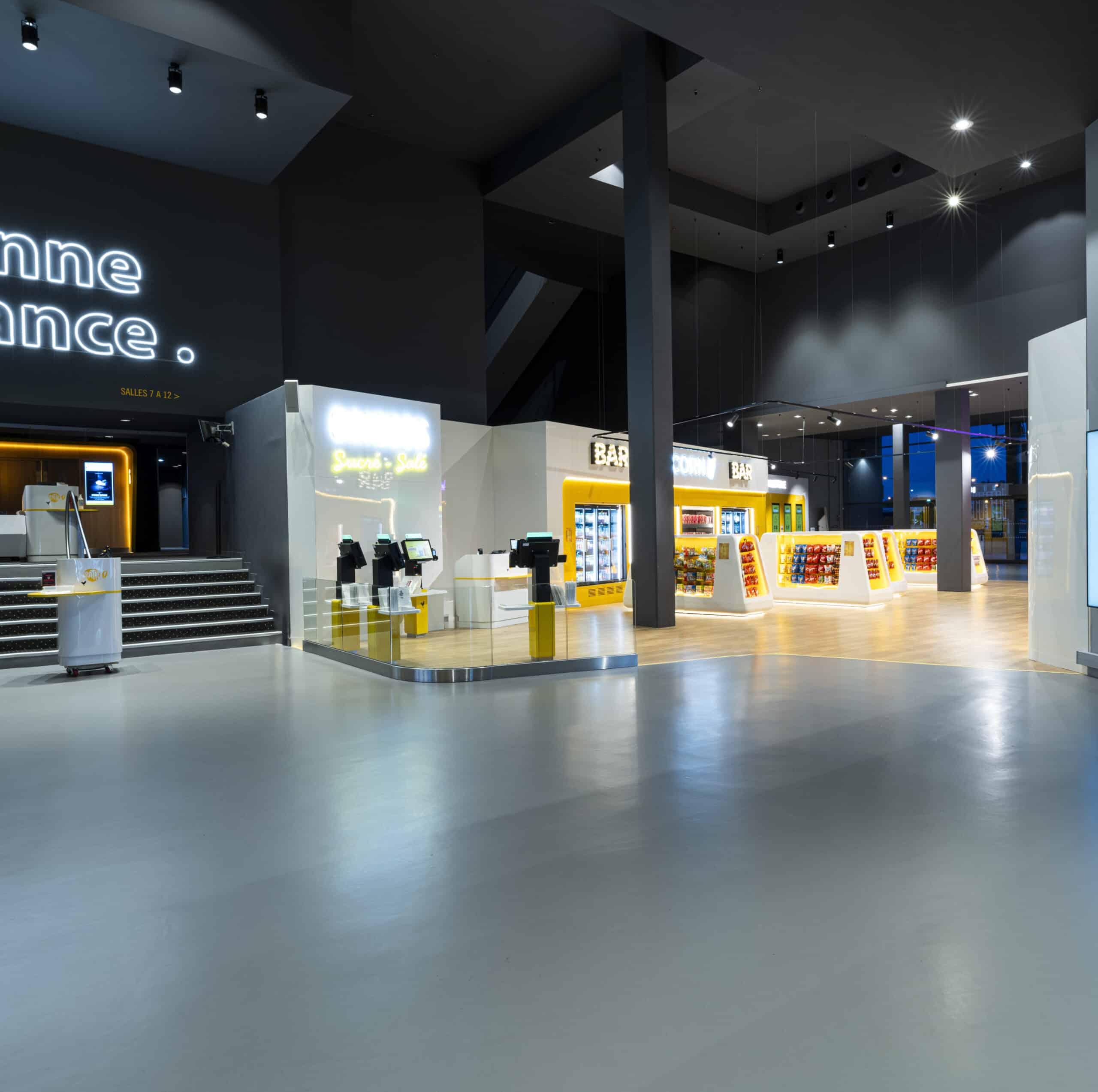 Hall de cinéma spacieux et moderne avec un bar lumineux proposant des confiseries, des bornes automatiques, et des indications vers les salles.
