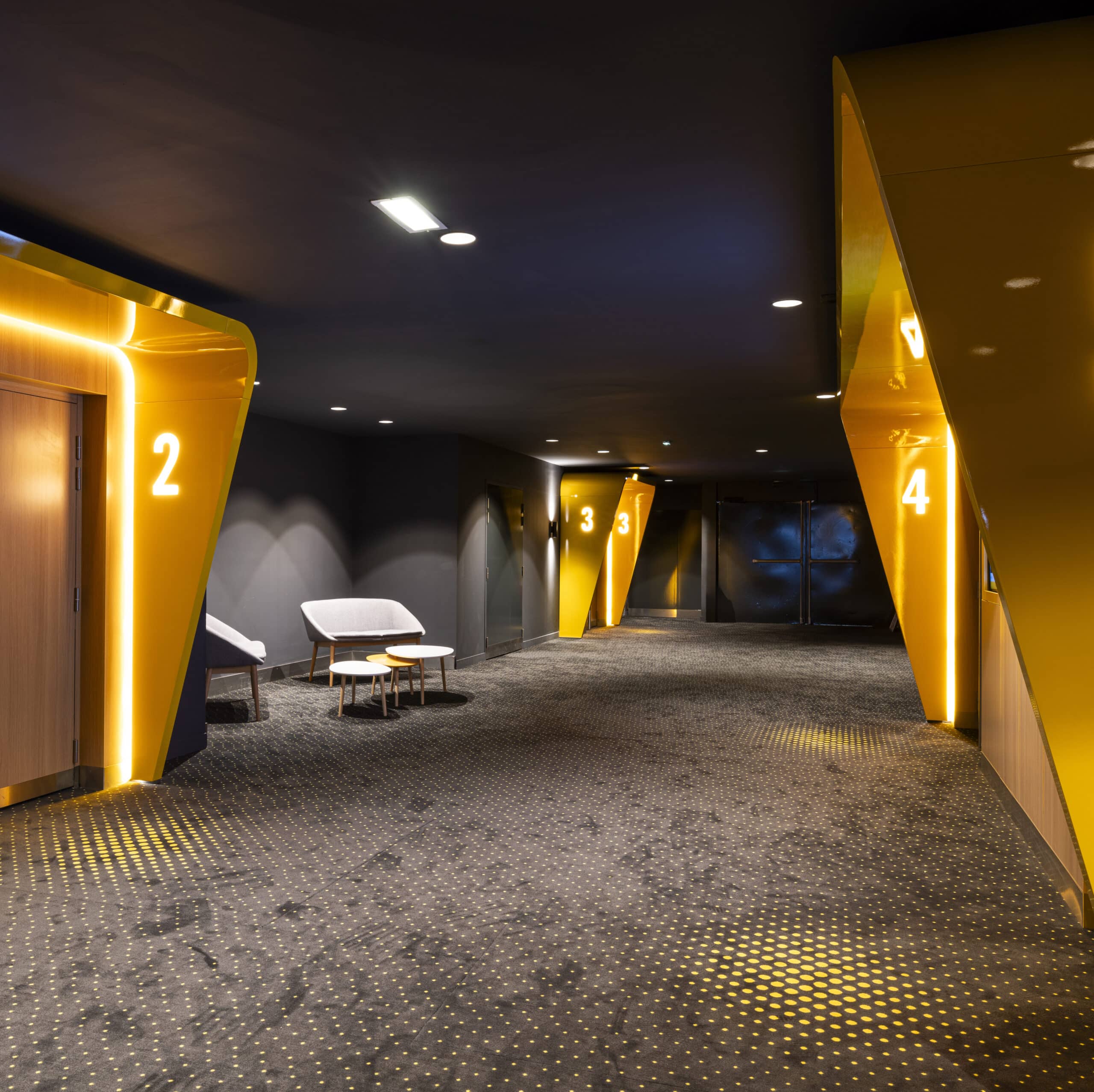 Couloir dans un cinéma avec signalisation lumineuse des salles 2 à 4, un tapis sombre à motifs, et des assises modernes.
